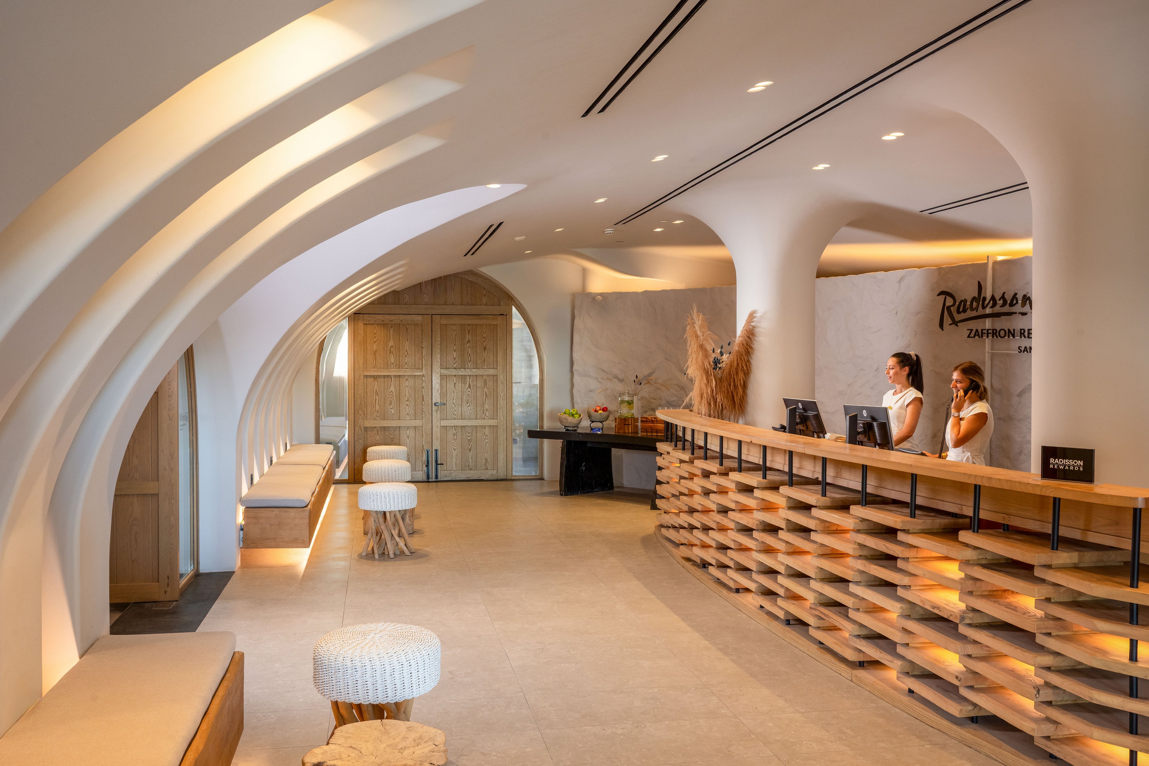 A contemporary hotel lobby features a long wooden reception desk staffed by two attendants. The space is defined by dramatic white curved arches and smooth columns that create a flowing, organic ceiling. Warm neutral tones, wooden doors, and textured stools contribute to a welcoming atmosphere. Soft lighting and minimalist decor emphasize the modern, stylish interior design.