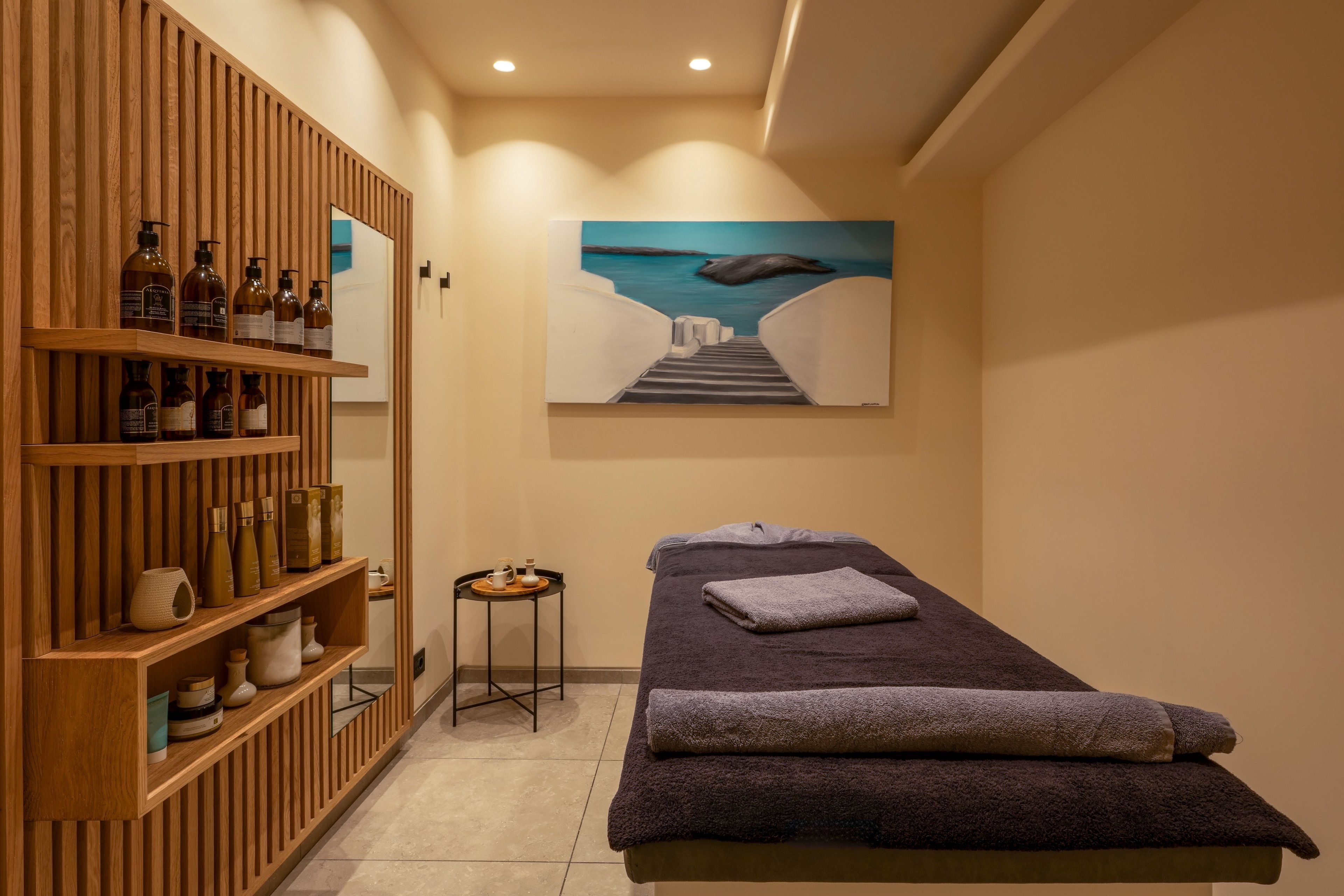 A neatly arranged spa treatment room features a massage bed with folded towels ready for use. Wooden shelves on the left display various bottles and wellness products, adding texture and warmth to the space. Soft, recessed ceiling lights create a calm, inviting atmosphere. A large wall painting of a coastal staircase view enhances the relaxing, retreat-like mood.