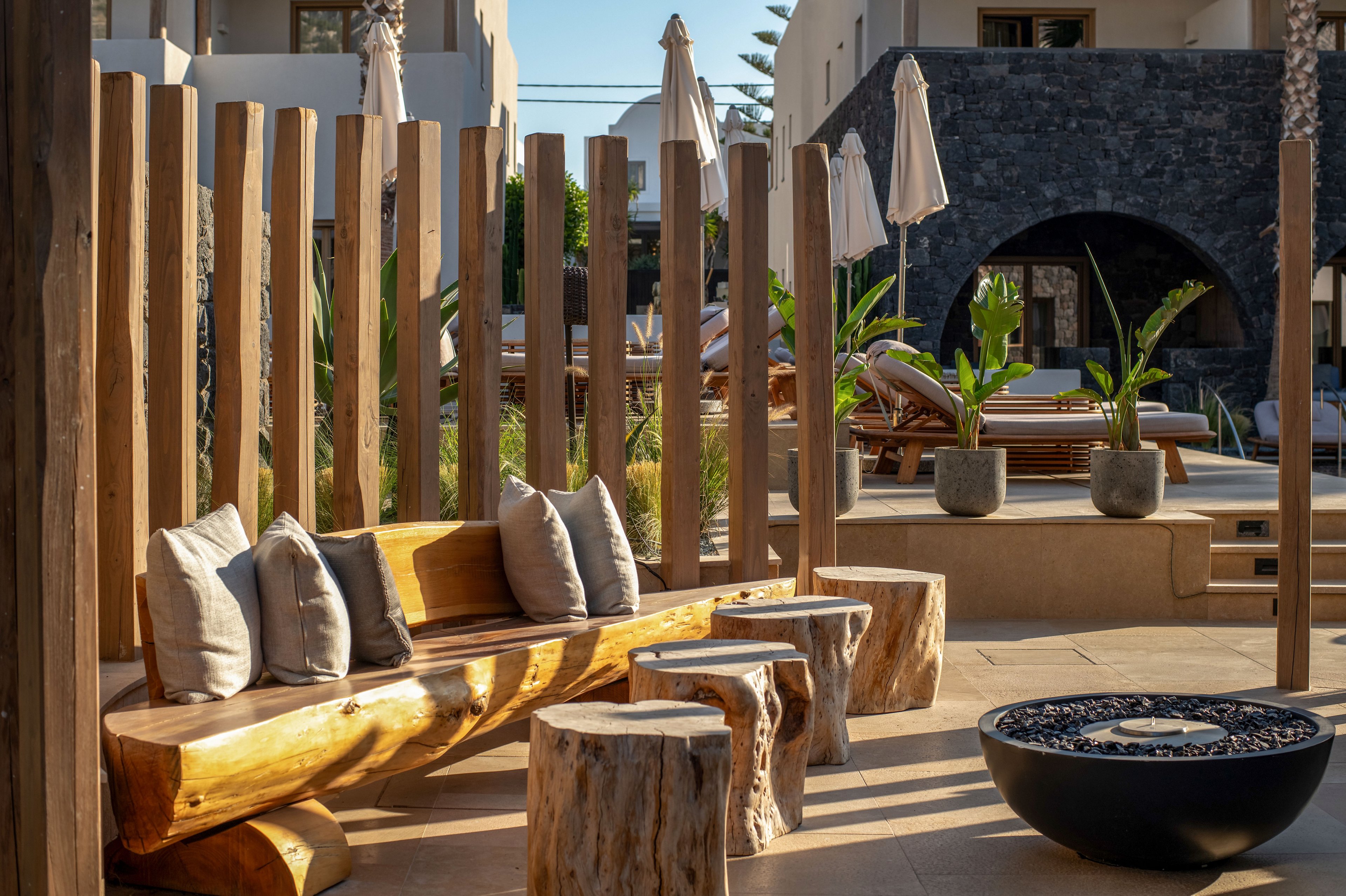 An inviting outdoor lounge area features a long rustic wooden bench with several cushions and matching log-style stools. Vertical wooden posts frame the seating space, creating a warm, natural atmosphere. In the background, contemporary resort buildings, potted tropical plants, and sun loungers with umbrellas suggest a relaxing vacation setting. A round fire pit or decorative bowl sits in the foreground, enhancing the cozy, modern design.