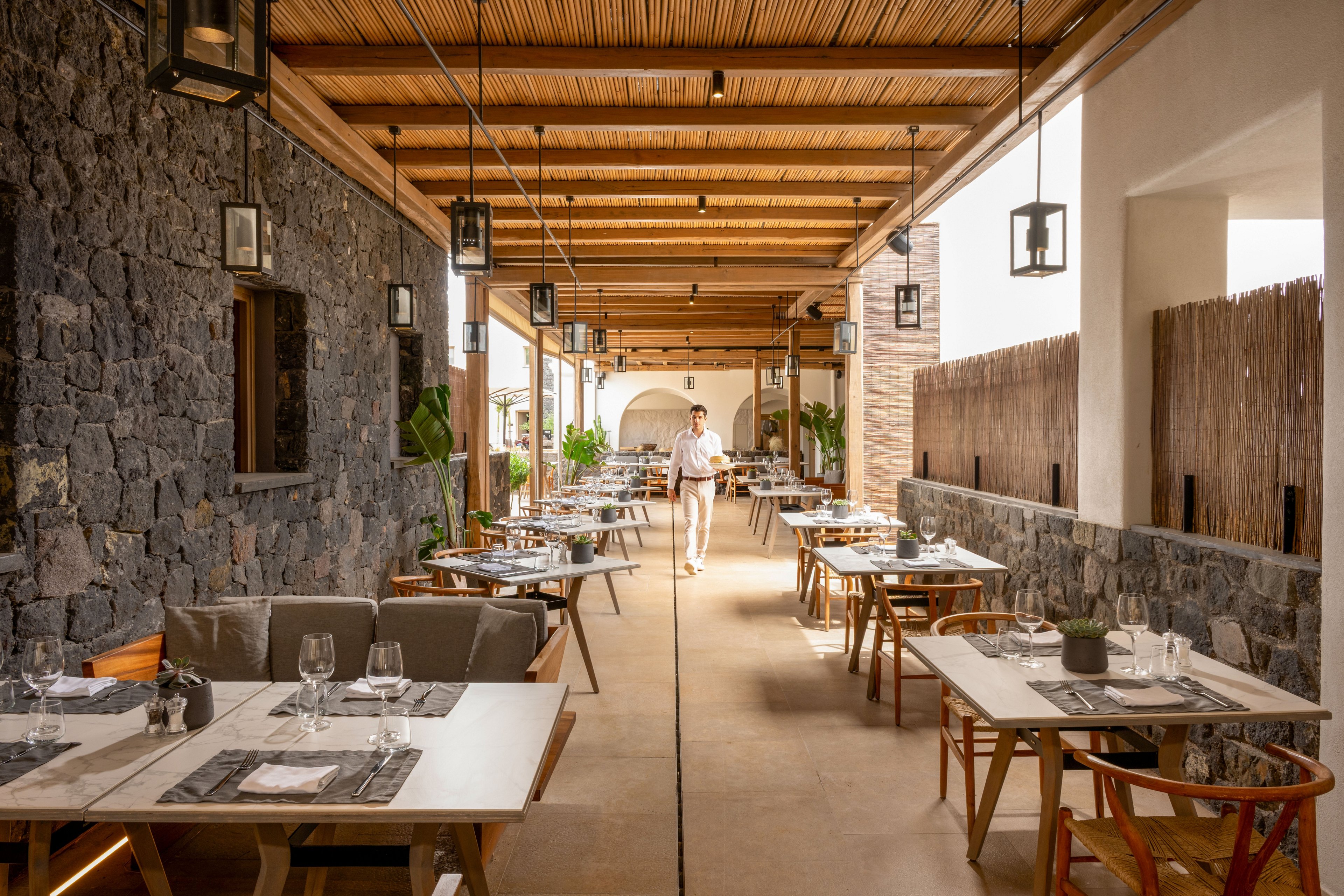 A bright open-air restaurant terrace features neatly arranged tables set with glassware and tableware. A waiter walks down the central aisle, adding a sense of service and hospitality to the scene. The space combines stone walls, wooden ceiling slats, and hanging lanterns for a warm, contemporary look. Natural light and potted plants enhance the inviting atmosphere of this dining area.