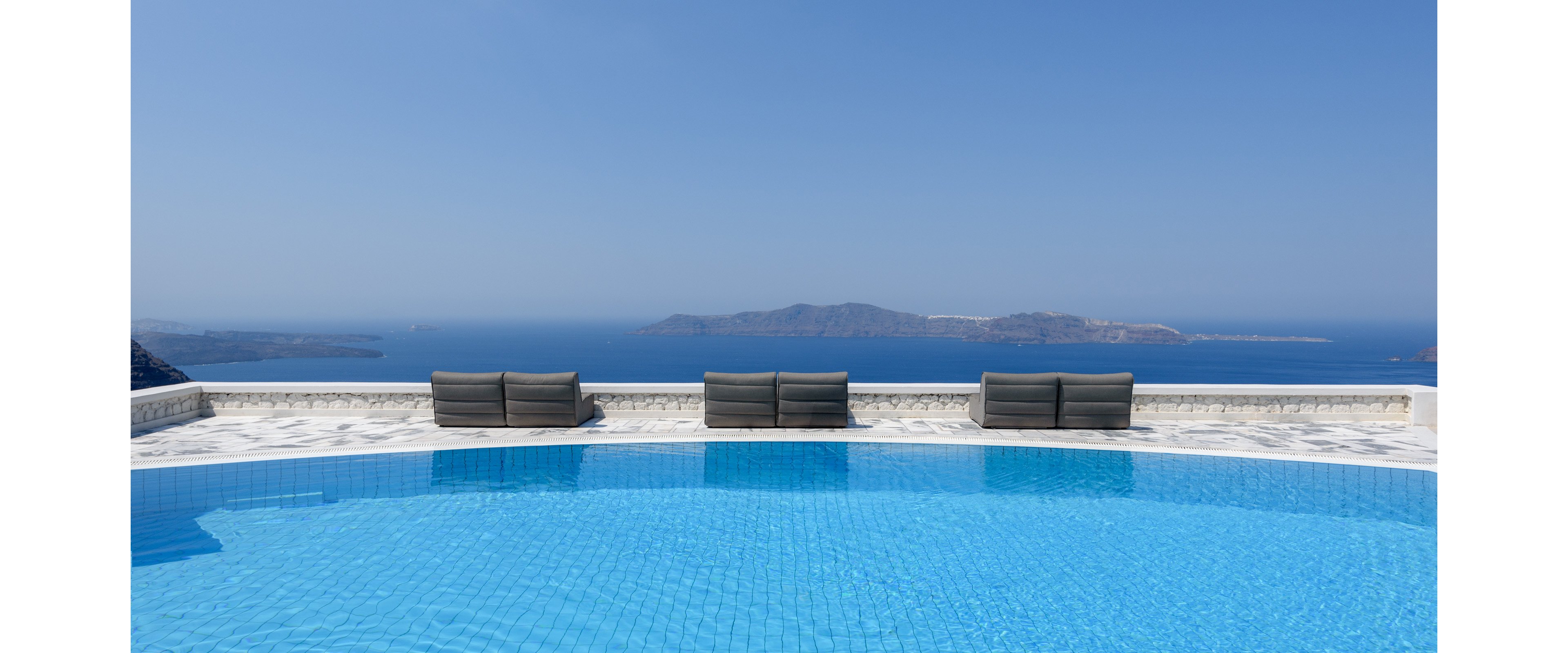 A woman in a white dress and sun hat stands at the edge of a bright blue infinity pool. She looks out over a vast seascape with distant islands under a clear blue sky. Modern outdoor lounge chairs line the poolside terrace, emphasizing a luxurious resort setting. The scene conveys calm, relaxation, and a serene summer vacation atmosphere.