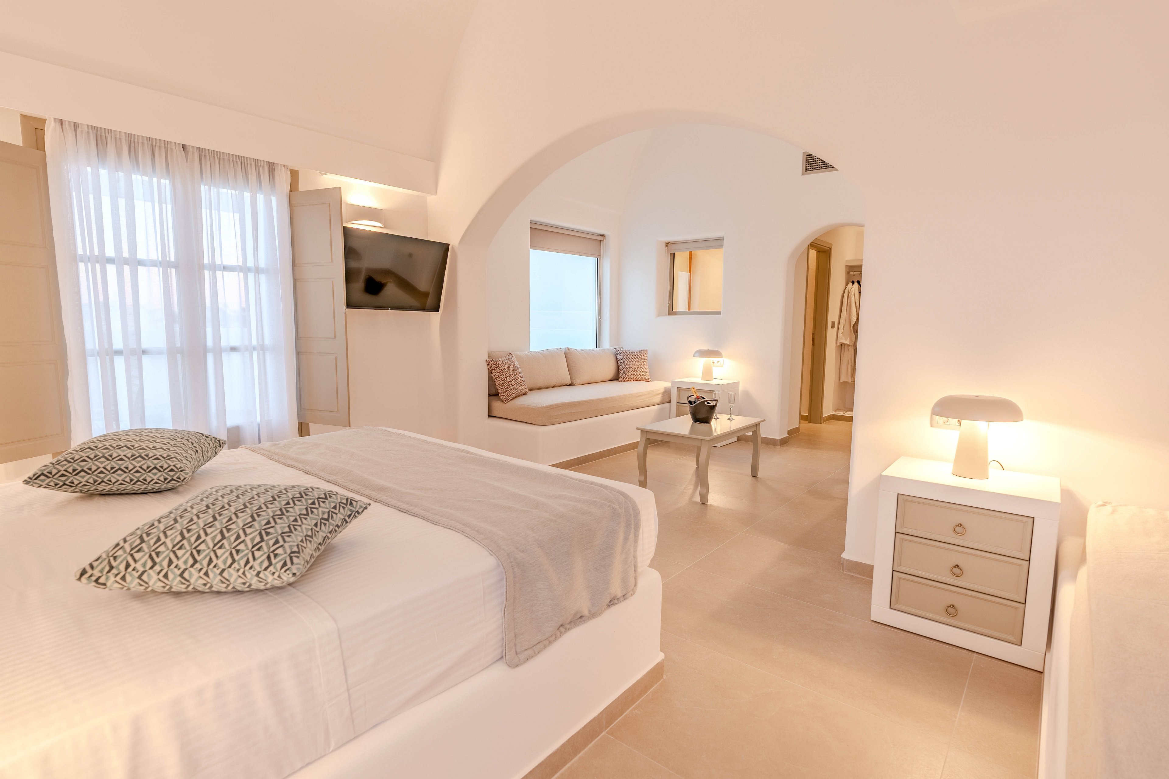 A softly lit hotel bedroom features a large bed with patterned pillows and a light gray throw. The room includes a wall-mounted television, a cozy built-in sofa, and a small table with decorative items. Warm white lighting from a modern bedside lamp and recessed fixtures enhances the neutral color palette. Arched doorways and sheer curtains contribute to the airy, serene atmosphere.