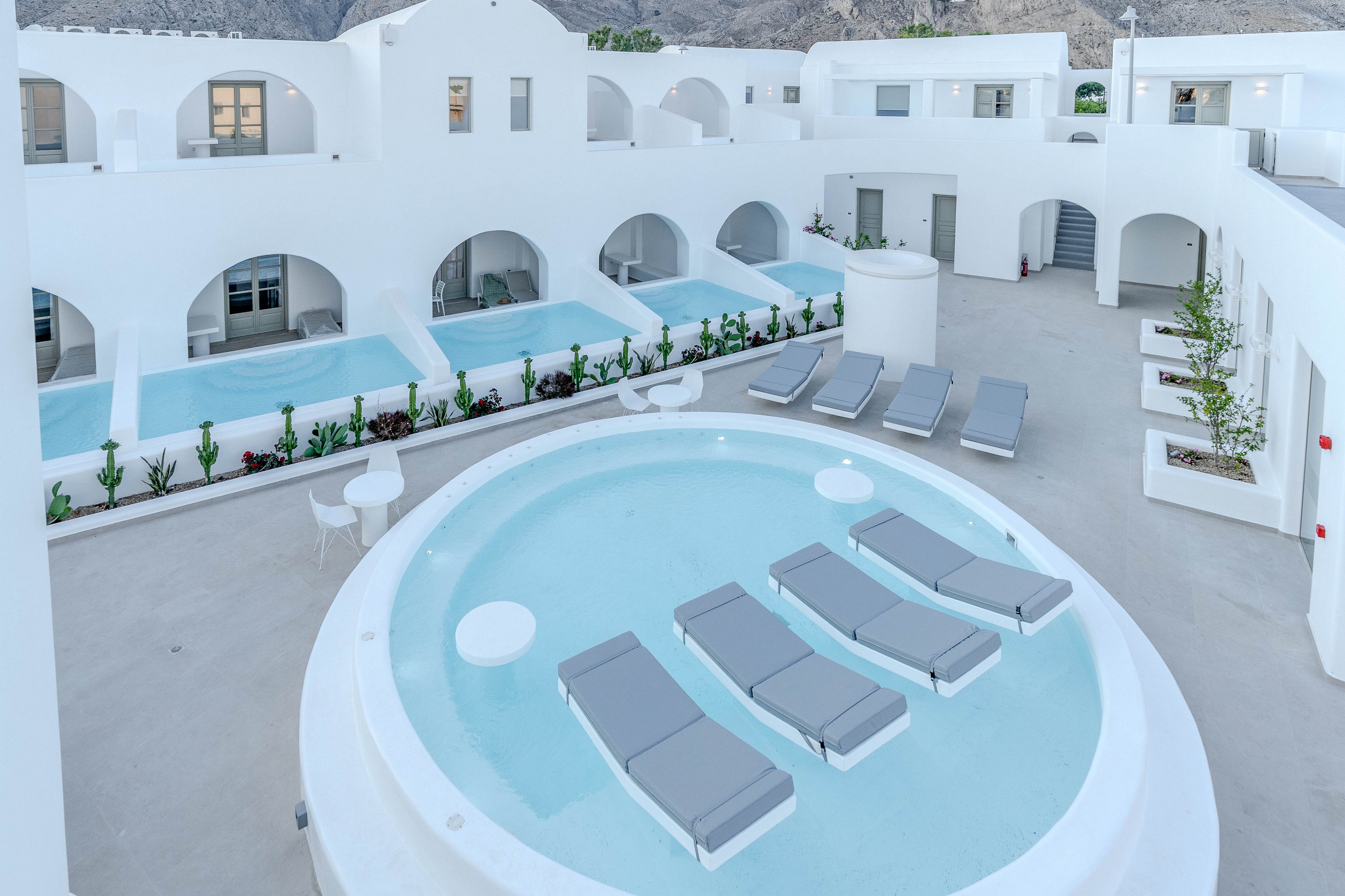 A contemporary white resort courtyard features a large circular pool with four gray cushioned loungers partially submerged in the water. Surrounding the central pool are several rectangular private pools aligned along the white building facades. Minimalist outdoor furniture and sun loungers are arranged on the smooth concrete terrace, creating a clean and relaxing atmosphere. The architecture is characterized by curved arches, multiple levels, and bright white surfaces that reflect the soft daylight.