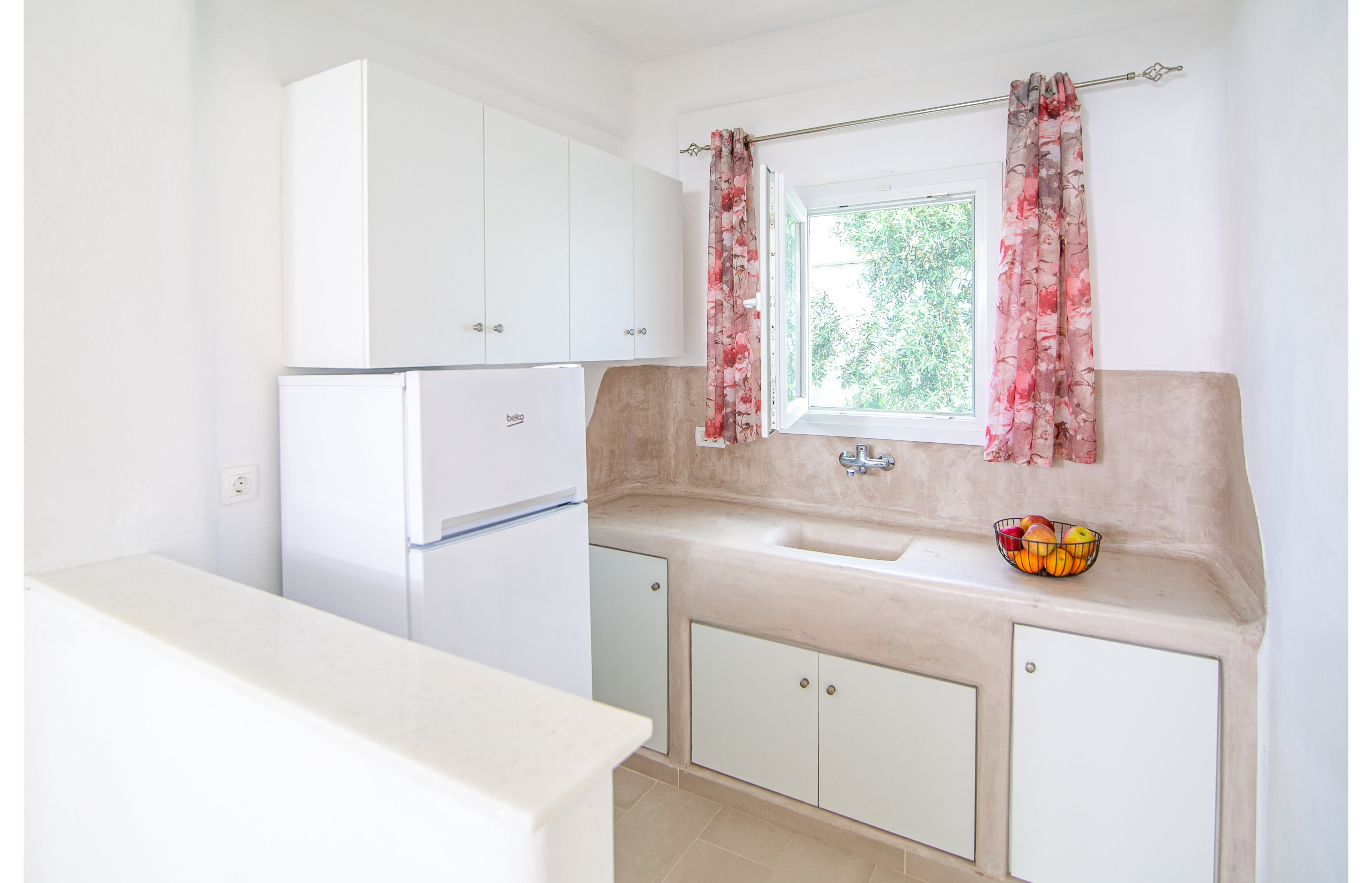 A compact kitchen featuring white cabinets and a white refrigerator. The setting includes a beige countertop and sink, with a bowl of colorful fruit adding vibrancy. Red and white patterned curtains frame the window, which offers a view of greenery outside.