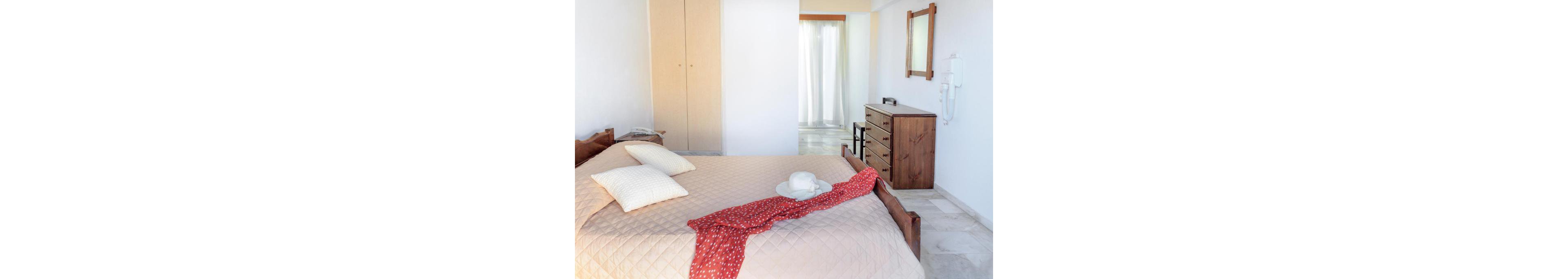A simple bedroom features a wooden bed with a beige quilt and two light-colored pillows. A white hat and red patterned scarf are placed casually on the bed, adding a touch of color. In the background, a wooden dresser, wall mirror, and built-in wardrobe complete the furnishings. Sheer curtains let in soft natural light, creating an airy and relaxed atmosphere.