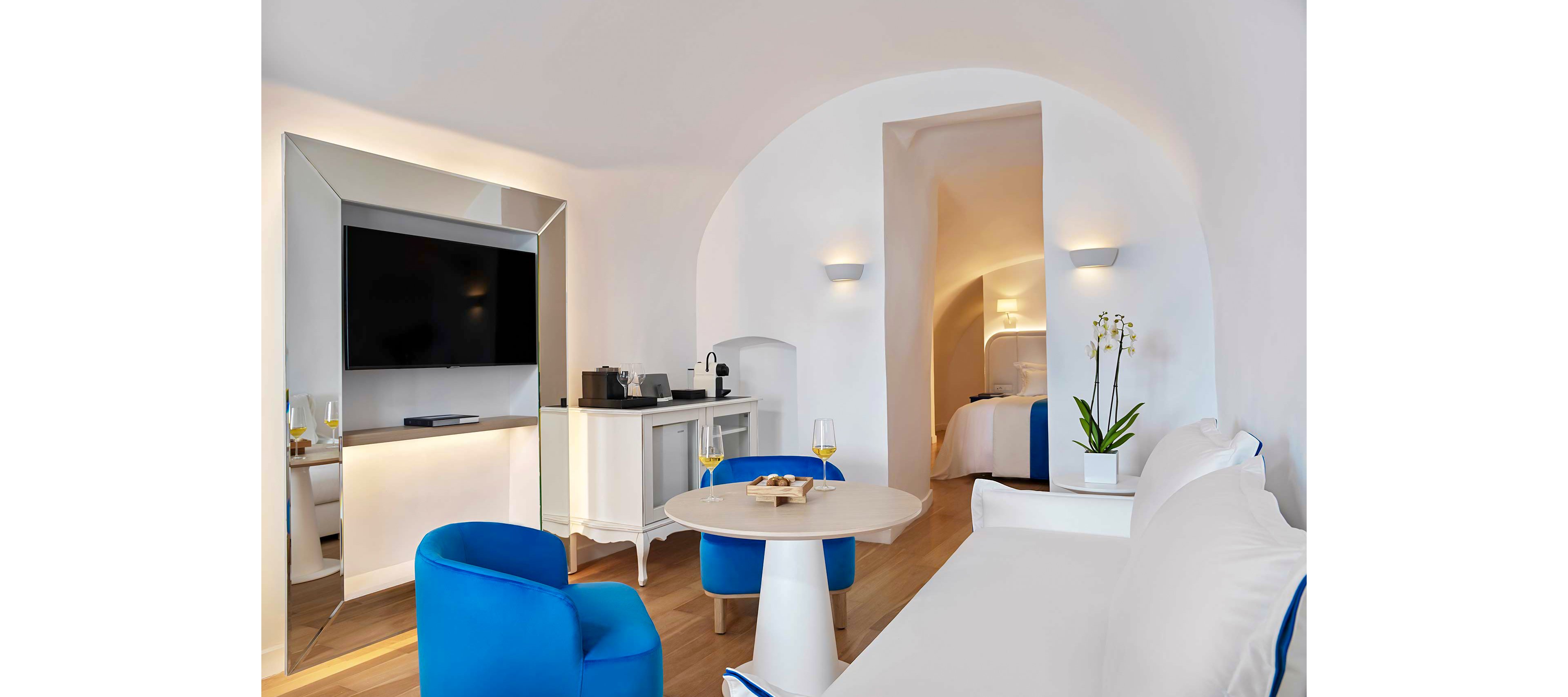 A stylish hotel suite featuring a sleek flat-screen TV, a cozy seating area with bright blue chairs, and a minimalist dining table. The setting includes a white and blue color palette, complemented by natural wood flooring and soft lighting. Visible details include a coffee machine and wine glasses on the table, creating a relaxed and luxurious atmosphere.