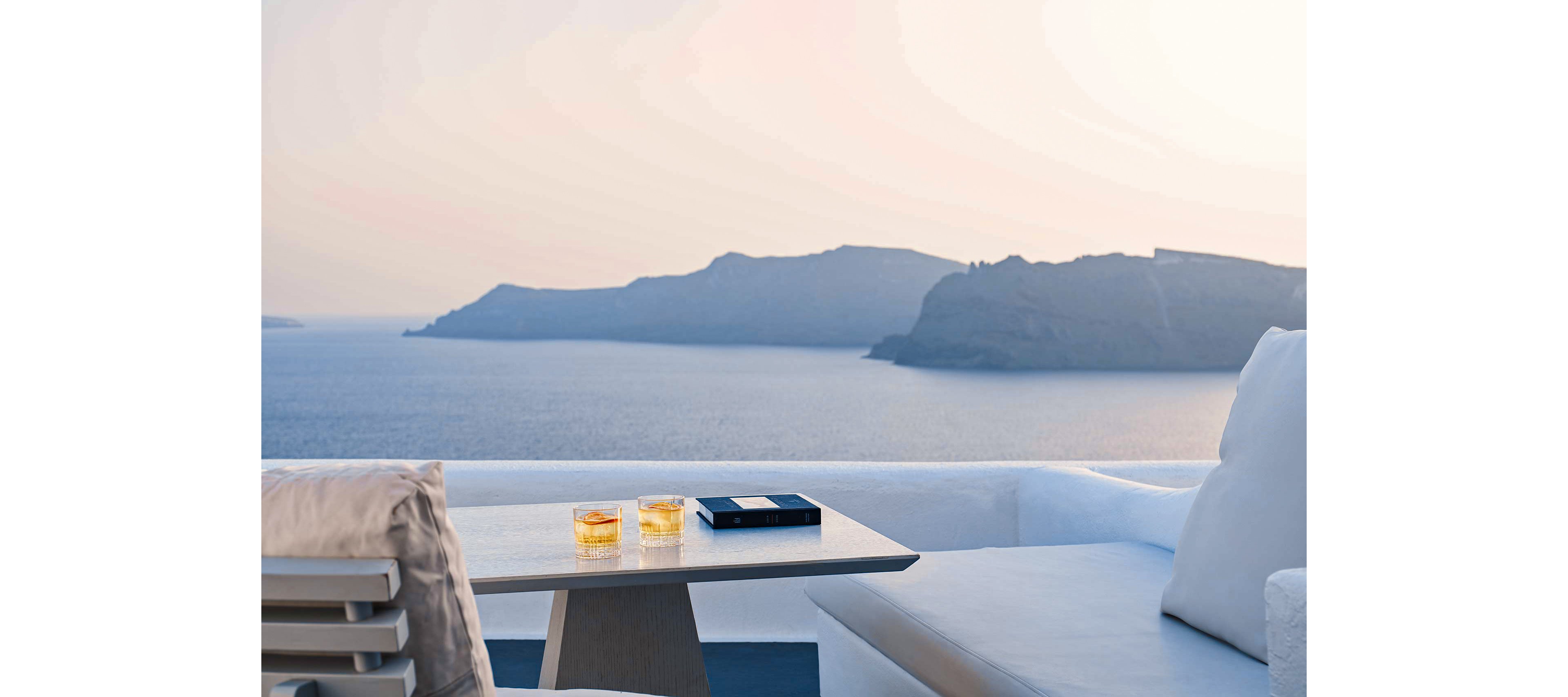 A serene coastal setting featuring a modern terrace overlooking the sea. Two glasses of amber-colored beverages sit on a table alongside a book, suggesting relaxation and leisure. The backdrop includes distant cliffs under a soft pastel sky, creating a tranquil atmosphere.