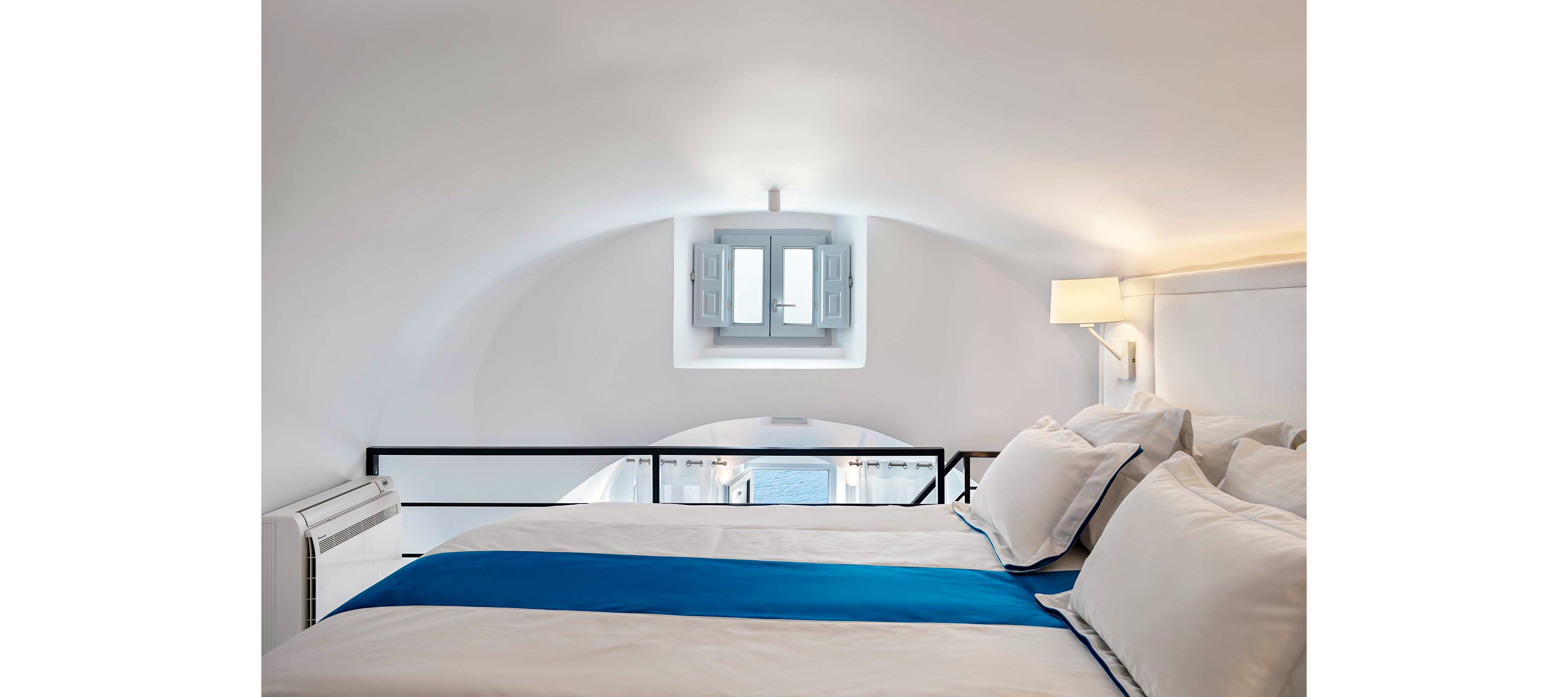 A cozy loft-style bedroom featuring a neatly made bed with white linens and a blue accent runner. The room is illuminated by soft lighting and natural light streaming through a small window with shutters. The window offers a serene view of the ocean, complementing the minimalist and airy design of the space.