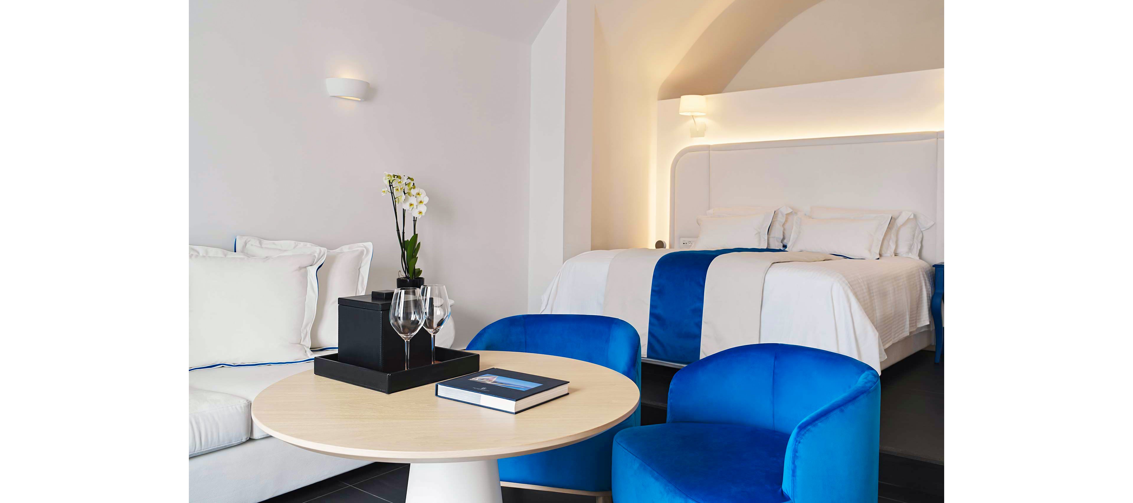 A stylish and modern hotel room featuring a neatly made bed with white and blue linens. The setting includes a cozy seating area with vibrant blue chairs and a round wooden table. Decorative elements such as a black tray with glasses and a white orchid add sophistication to the space. The room is illuminated by soft, warm lighting, creating a serene atmosphere.