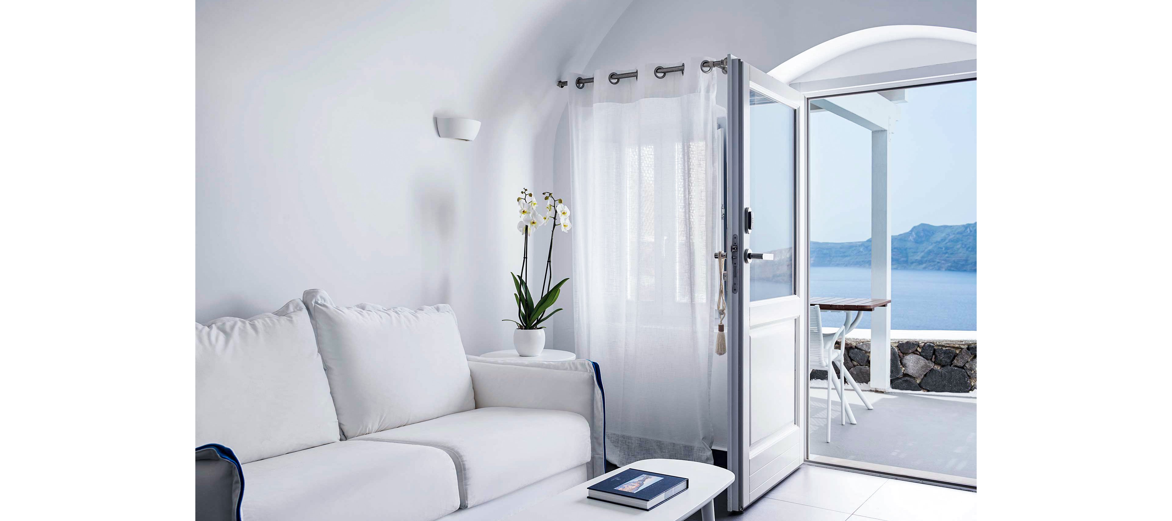 A serene white living room featuring a plush sofa and a potted orchid on a side table. The open door leads to a stunning ocean view with clear blue skies and a stone patio. The minimalist decor emphasizes tranquility and coastal elegance.