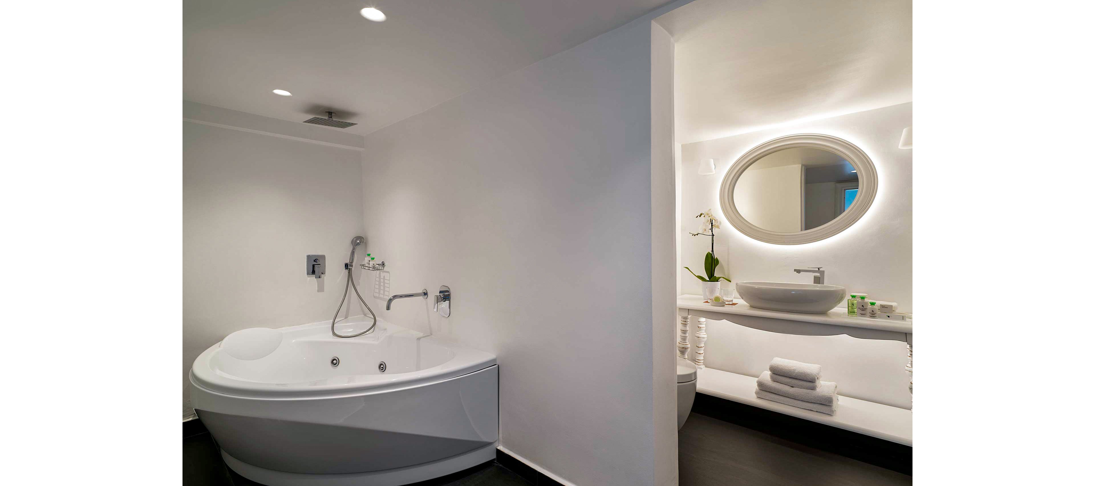 A sleek, contemporary bathroom featuring a white oval bathtub with chrome fixtures. Adjacent is a vanity area with a round illuminated mirror, a white sink, and neatly folded towels. The setting is minimalistic with soft lighting and a clean aesthetic.
