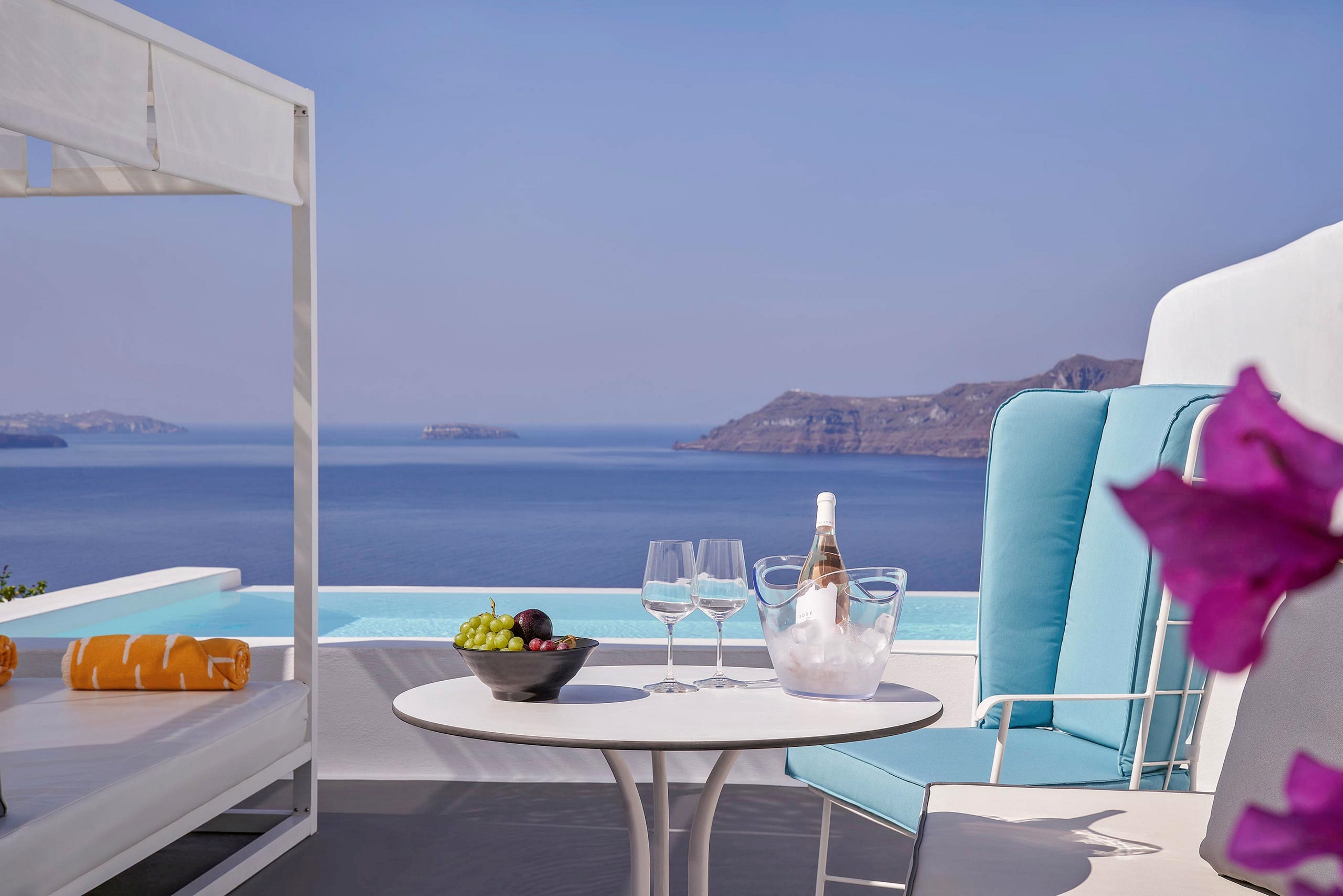 A stylish terrace overlooks a calm blue sea and distant islands. In the foreground, a round white table holds a bucket of ice with a champagne bottle, three empty wine glasses, and a plate of fresh grapes. Cushioned turquoise chairs and a white sunbed with an orange towel suggest a relaxing vacation setting. Bright sunlight and clear skies create a serene, luxurious holiday atmosphere with vibrant pops of color from nearby flowers.