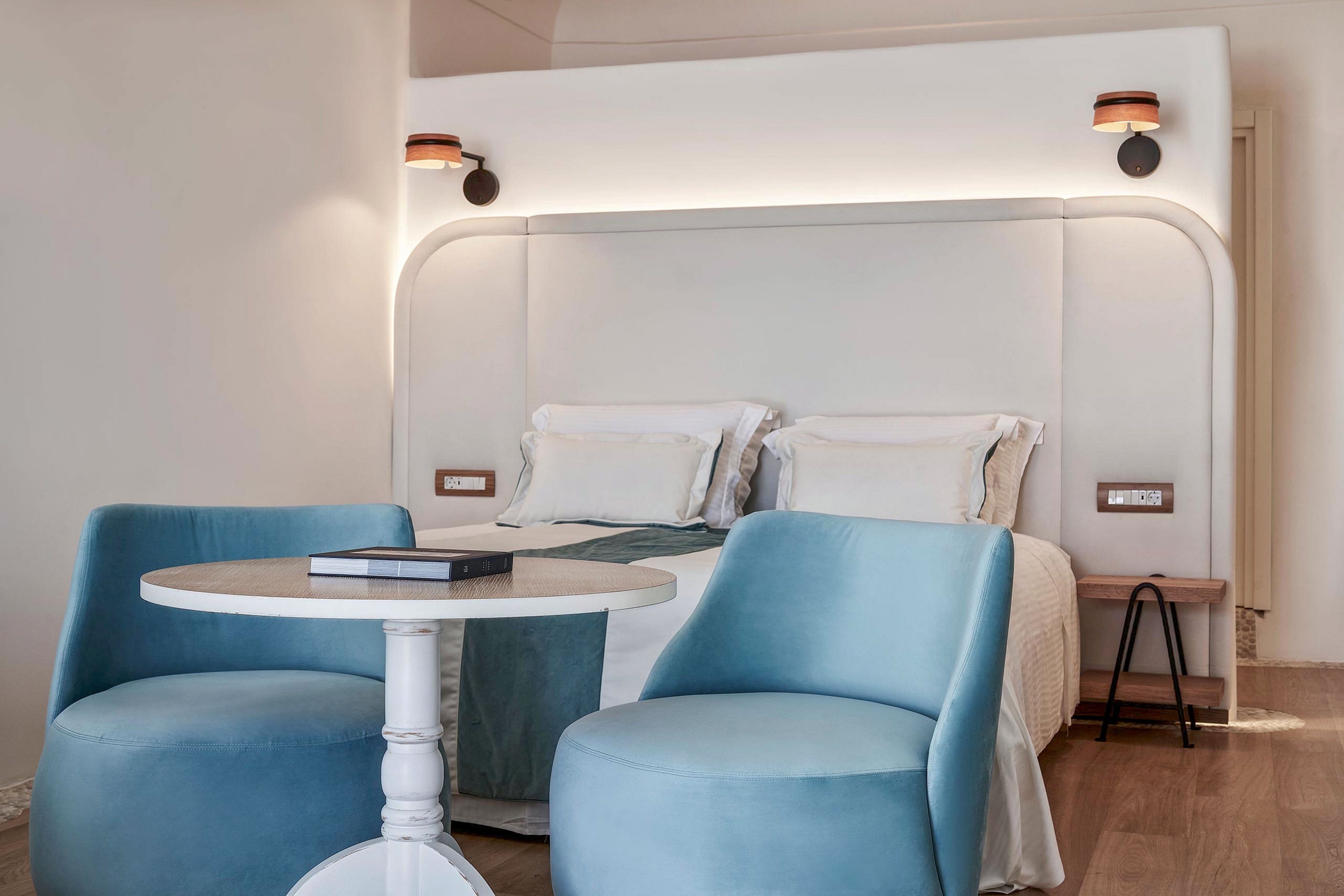 A contemporary hotel bedroom features a neatly made double bed with white linens and soft teal accents. In the foreground, two plush light blue chairs surround a small round white table with a closed book on top. The room is illuminated by warm wall sconces above the oversized upholstered headboard. Minimalist decor and neutral walls create a calm, inviting atmosphere suitable for relaxation or reading.