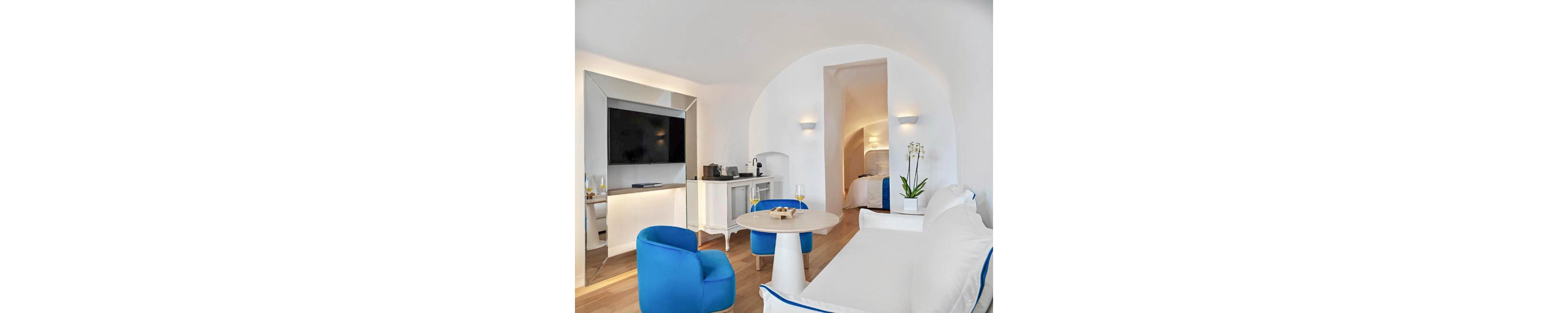 Bright contemporary hotel suite features white walls and furniture with bold blue chairs and trim. A flat-screen television is mounted above a console with a coffee machine and glassware. In the foreground, a round table holds two glasses of white wine and snacks beside a white sofa. An arched doorway leads to a cozy bedroom area with soft lighting and a neatly made bed. The space feels airy and minimalist with natural wood flooring and a potted orchid adding a touch of greenery.