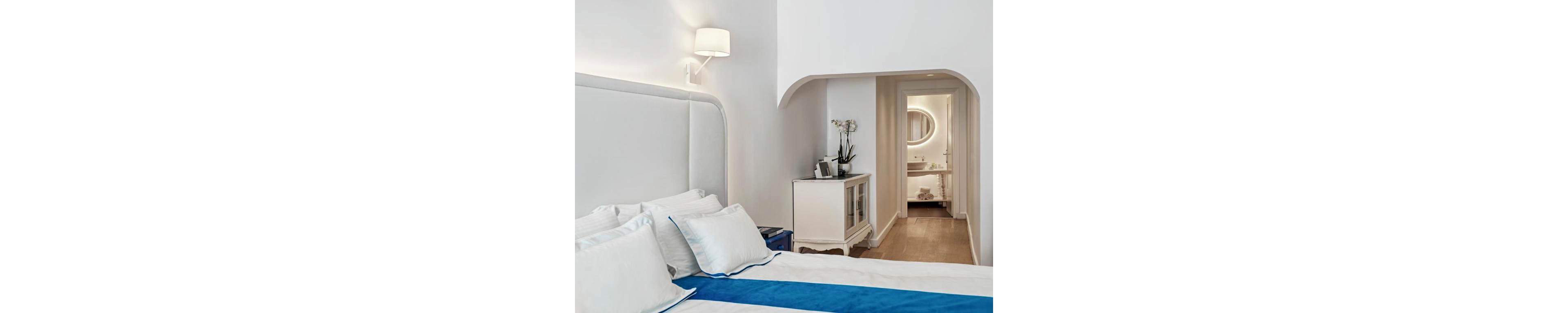 A modern hotel bedroom features a neatly made bed with white linens and a bold blue runner. The room is decorated in a minimalist style with white walls, a padded headboard, and a wall-mounted lamp. A small cabinet with decorative items and an orchid stands near an arched doorway leading to a brightly lit bathroom. Warm wooden flooring and soft ambient lighting create a calm, inviting atmosphere.