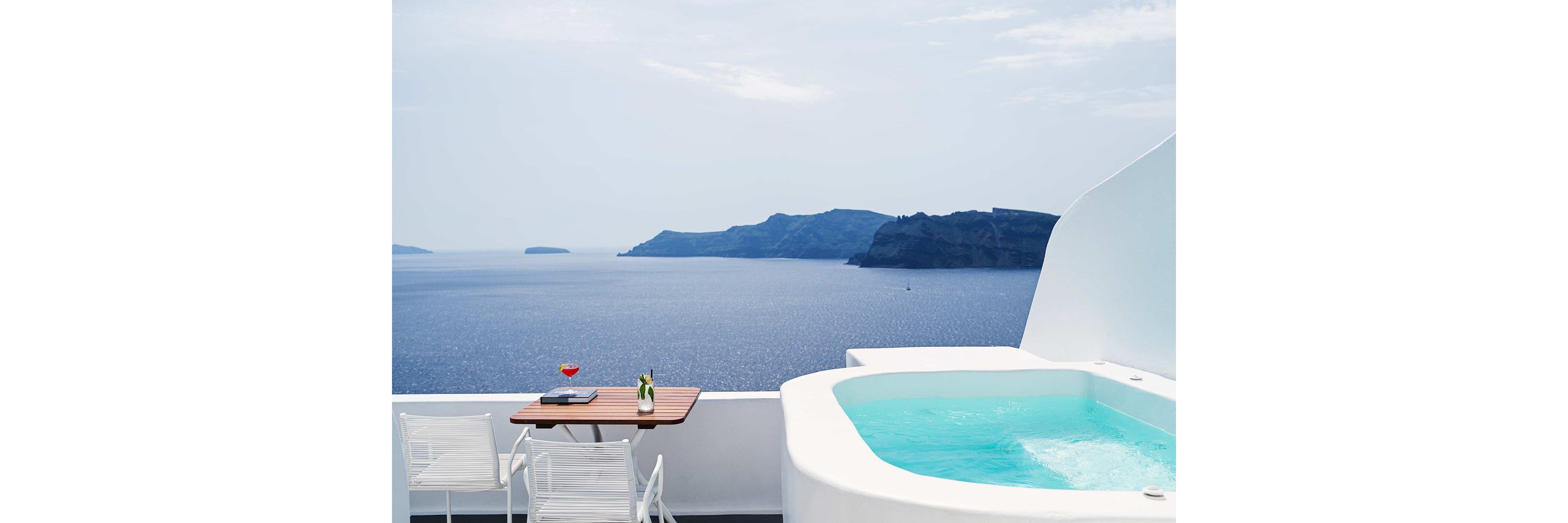 A small infinity pool sits on a white terrace with a sweeping view of a calm blue sea and distant islands. Beside the pool, a wooden table holds a laptop, a drink, and a bottle, with two white chairs arranged for relaxation. The scene features minimalist white architecture contrasting with the deep blue water and pale sky. Overall, the image evokes a serene, luxurious vacation atmosphere with a focus on leisure and quiet contemplation.