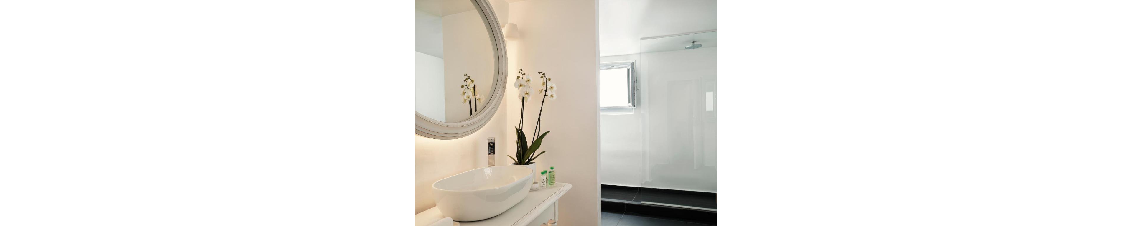 A contemporary bathroom features a sleek white vessel sink on a narrow countertop. A round wall mirror and soft lighting create a clean, minimalist look. White orchid flowers in a vase add a natural accent beside small toiletry bottles. In the background, a spacious walk-in shower with a glass partition and open window completes the bright, airy space.