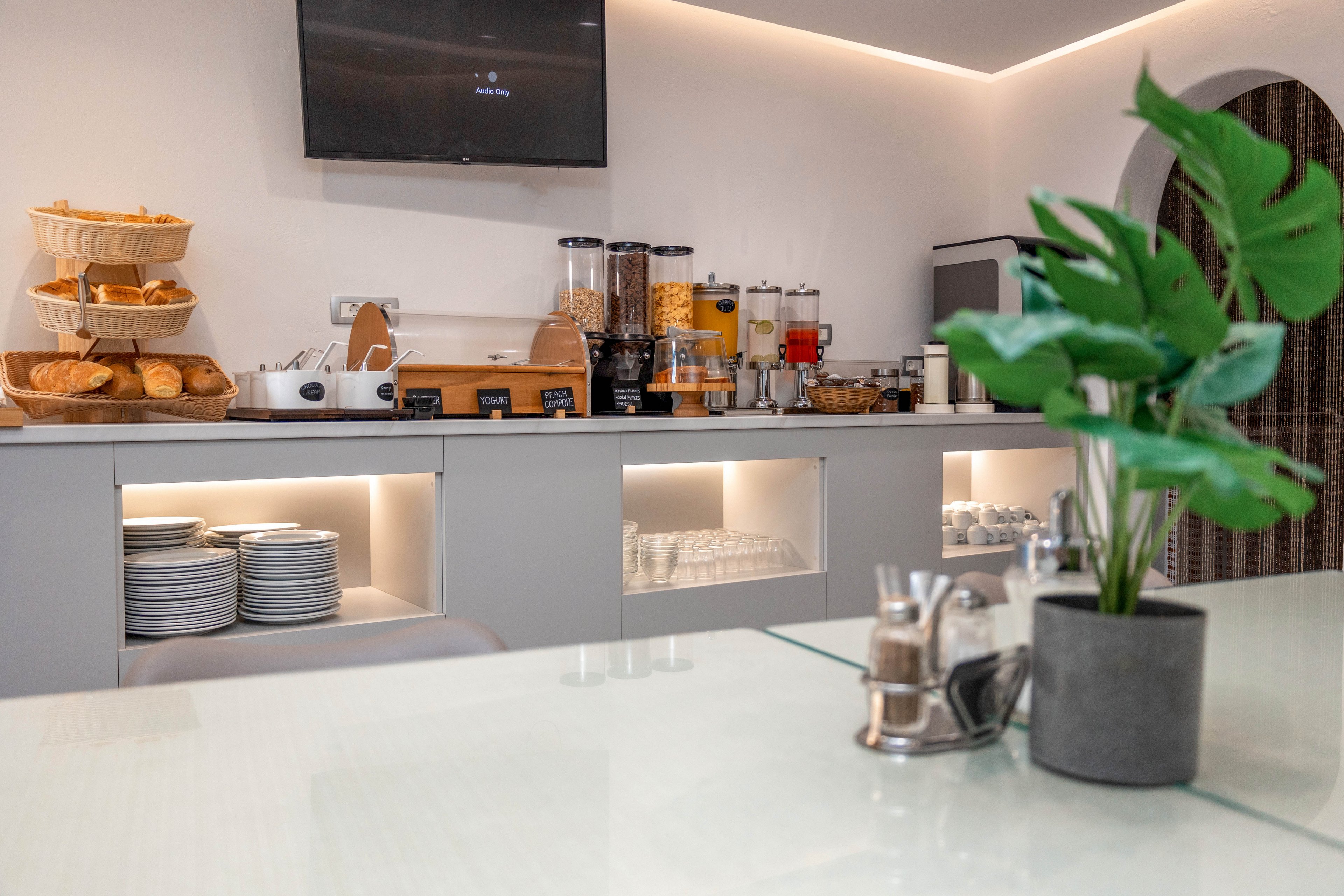 A contemporary breakfast buffet counter is set up with cereals, juices, and baked goods. Dispensers for cereal and juice stand beside baskets of bread and pastries, with stacks of plates neatly arranged below. Soft under-cabinet lighting and a wall-mounted television create a clean, modern ambiance. A small potted plant and condiments sit on the foreground counter, adding a touch of greenery to the minimalist space.