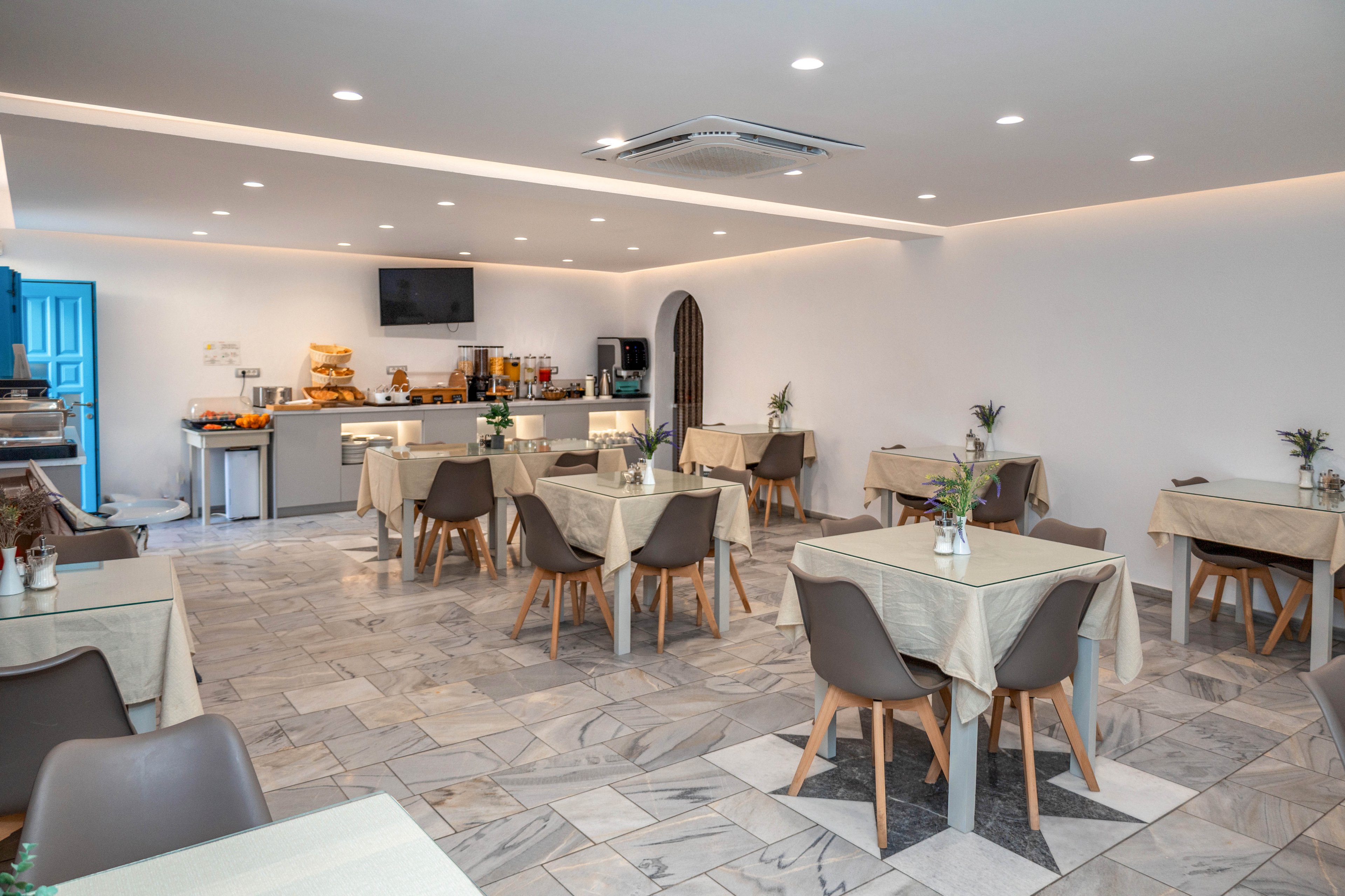 Contemporary dining room with neatly arranged tables and chairs is shown in a hotel or guesthouse setting. A self-service buffet counter with coffee machines, juice dispensers, and food trays is visible at the back. The space features neutral colors, tiled flooring, and recessed ceiling lighting for a clean, minimalist look. No people are present, emphasizing the tidy and organized atmosphere.
