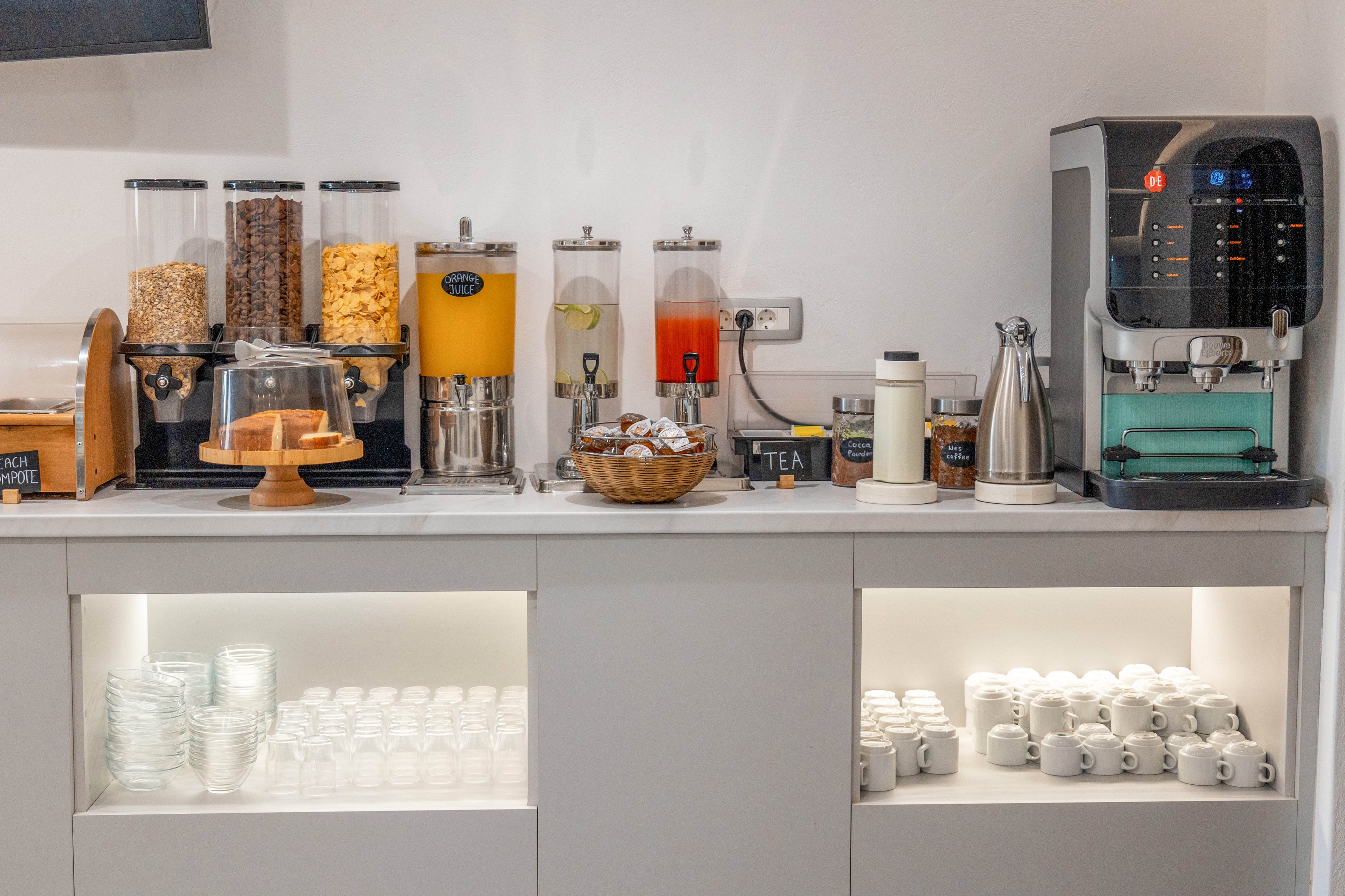A modern self-service breakfast station features cereal dispensers, juice containers, and a hot drink machine on a white counter. A cake stand with pastries and a basket of wrapped treats sit beside labeled jars for coffee and tea. Below the counter, shelves are neatly stocked with glass cups and white mugs. The overall setup suggests a clean, organized refreshment area in a hotel or office lounge.