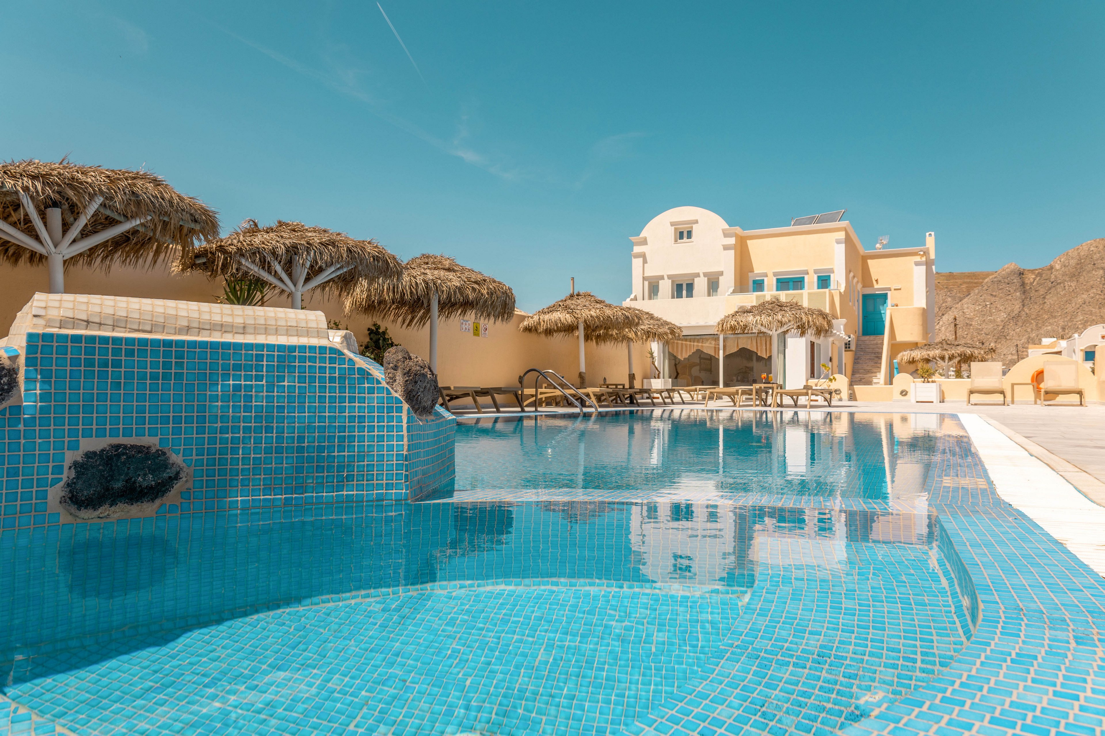 A bright blue tiled swimming pool dominates the foreground of a sunny resort scene. In the background, a light-colored villa-style building with arches and balconies stands against a clear blue sky. Straw parasols and empty sun loungers line the poolside, suggesting a relaxing vacation atmosphere. The warm tones of the architecture contrast with the cool turquoise water, evoking a tranquil Mediterranean or island getaway.
