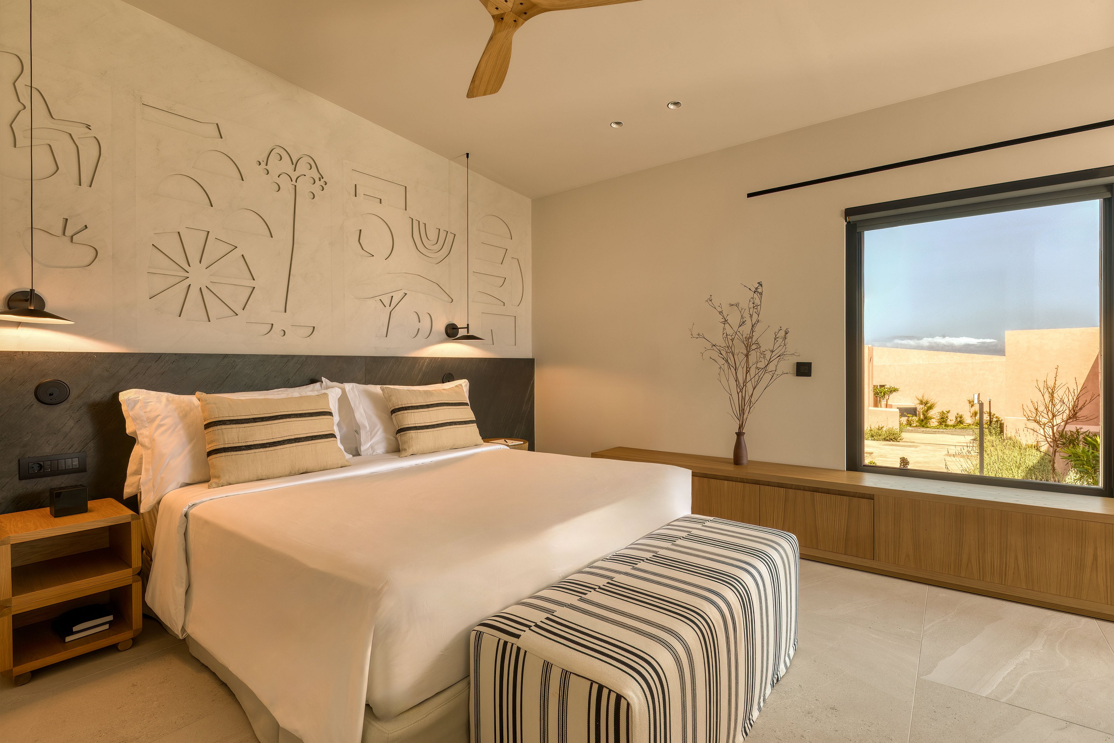 A contemporary hotel bedroom features a neatly made double bed with white linens and striped pillows. A matching striped bench sits at the foot of the bed, complementing the neutral tones of the room. Artistic wall carvings and pendant lights add visual interest above the headboard. A large window reveals an outdoor garden and distant landscape, bringing natural light into the calm, minimalist interior.