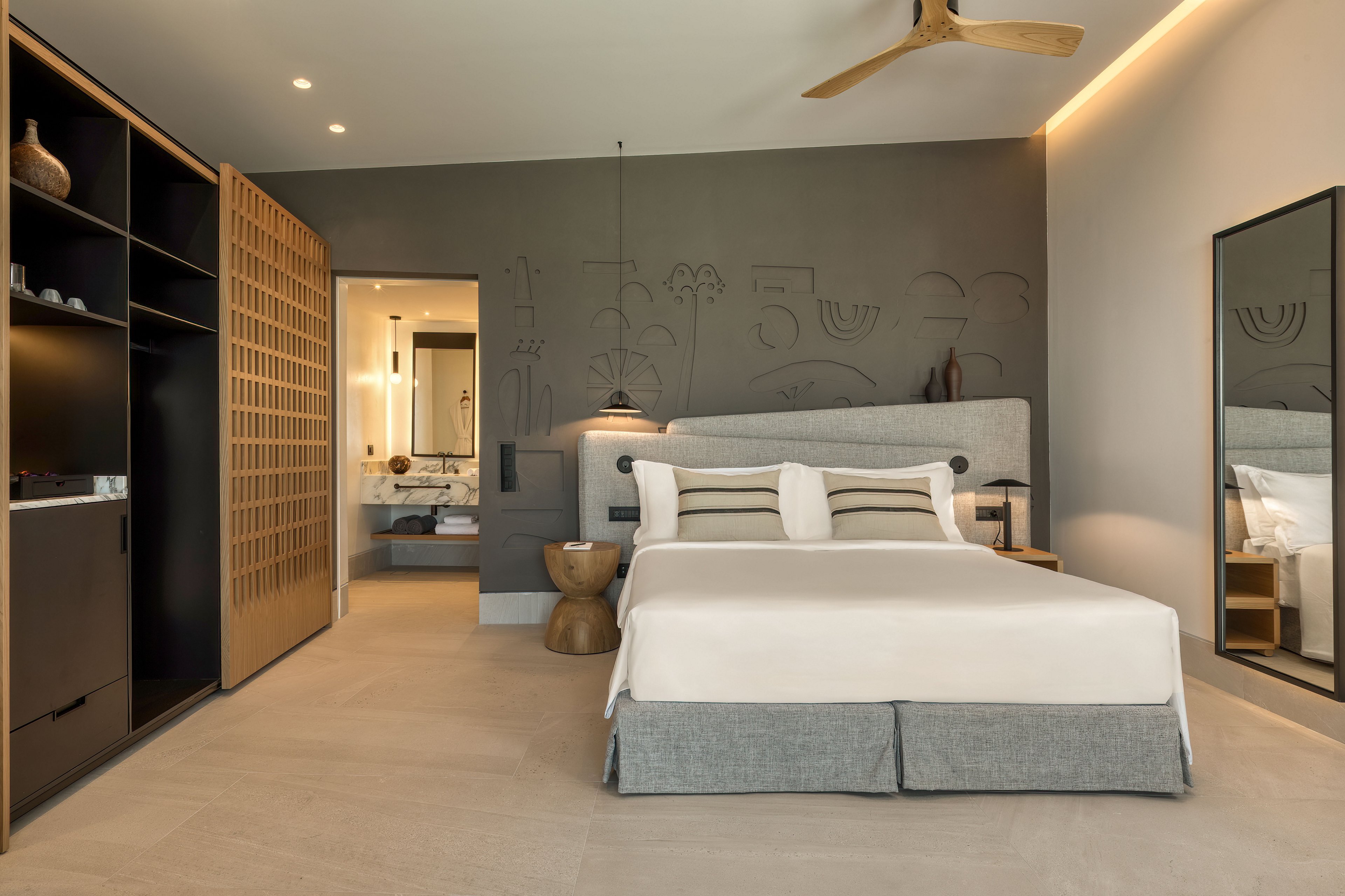 A contemporary bedroom features a large bed with neutral linens and a padded headboard against a dark accent wall. Built-in shelving and a sliding wooden screen frame the left side of the room, while a full-length mirror stands on the right. Warm lighting, a wooden ceiling fan, and sculptural bedside tables create a calm, minimalist atmosphere. An open doorway reveals an adjoining bathroom with a vanity and decorative objects, enhancing the sense of a stylish hotel suite.