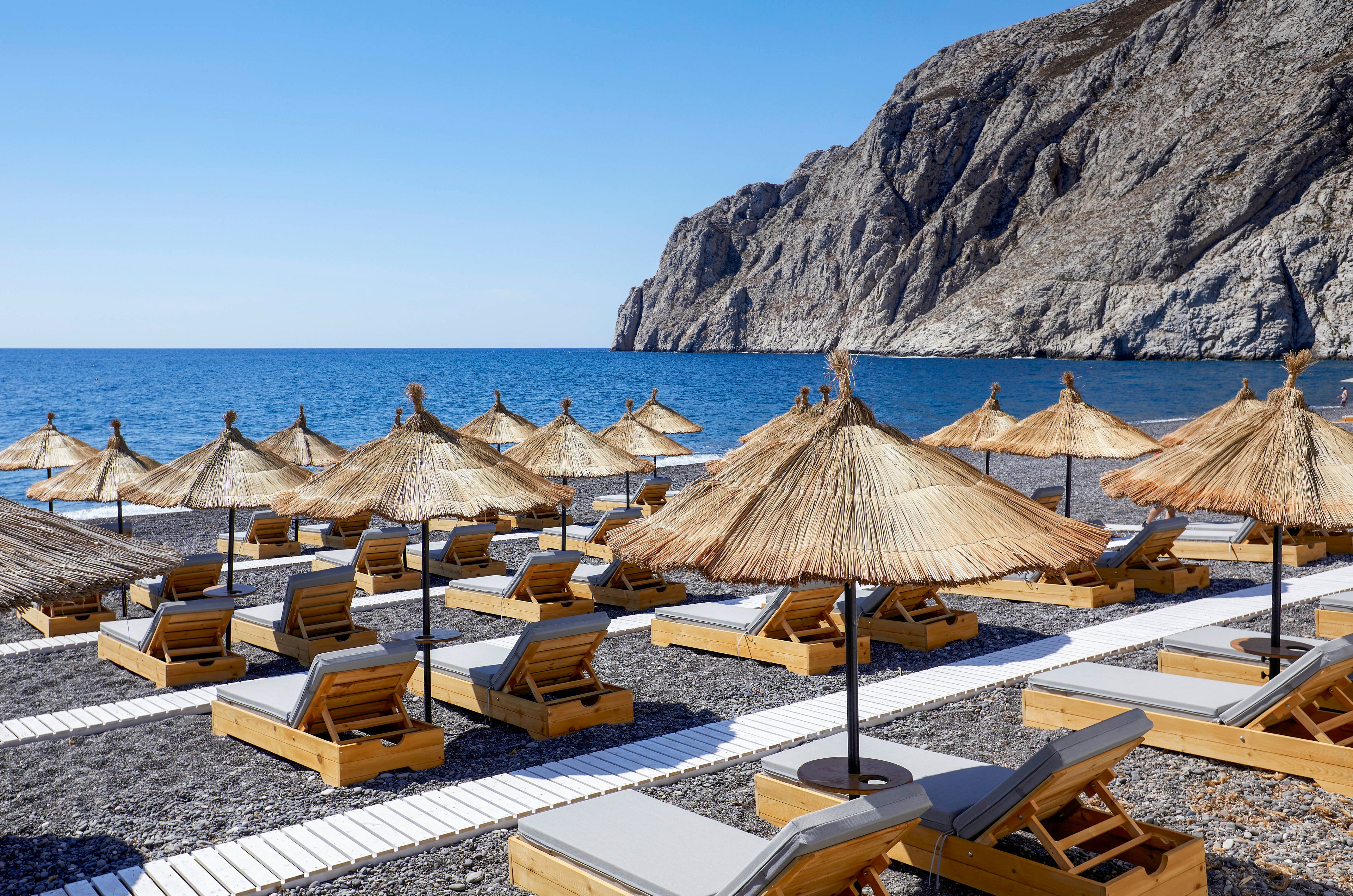 A row of wooden sun loungers with cushions is arranged neatly on a pebble beach under straw umbrellas. The beach faces a calm blue sea with gentle waves. In the background, steep rocky cliffs rise dramatically from the shoreline under a clear sky. The scene suggests a peaceful, uncrowded coastal resort ready for visitors.