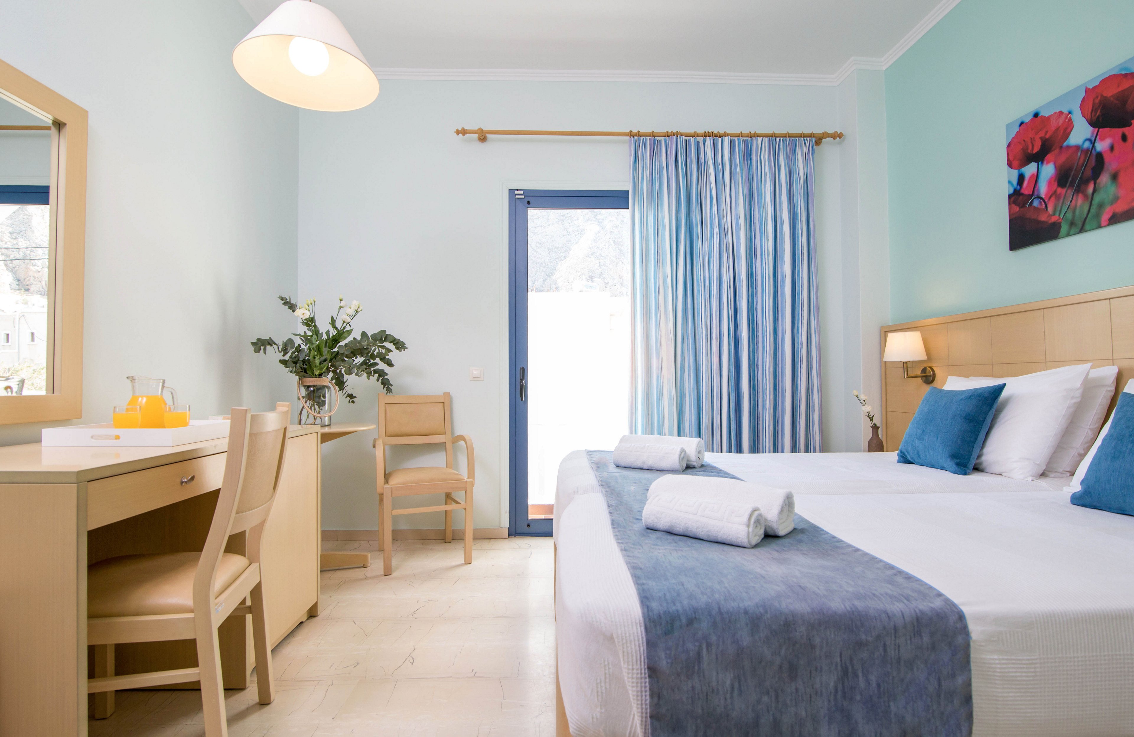 A neatly made double bed with white linens and blue accents stands in a bright, modern bedroom. The room features a wooden desk with a mirror, chair, and a tray with drinks beside a vase of flowers. Light blue walls, striped curtains, and a large window or door allow natural light to fill the space. A wall-mounted lamp and colorful artwork above the bed add warmth and decorative interest.