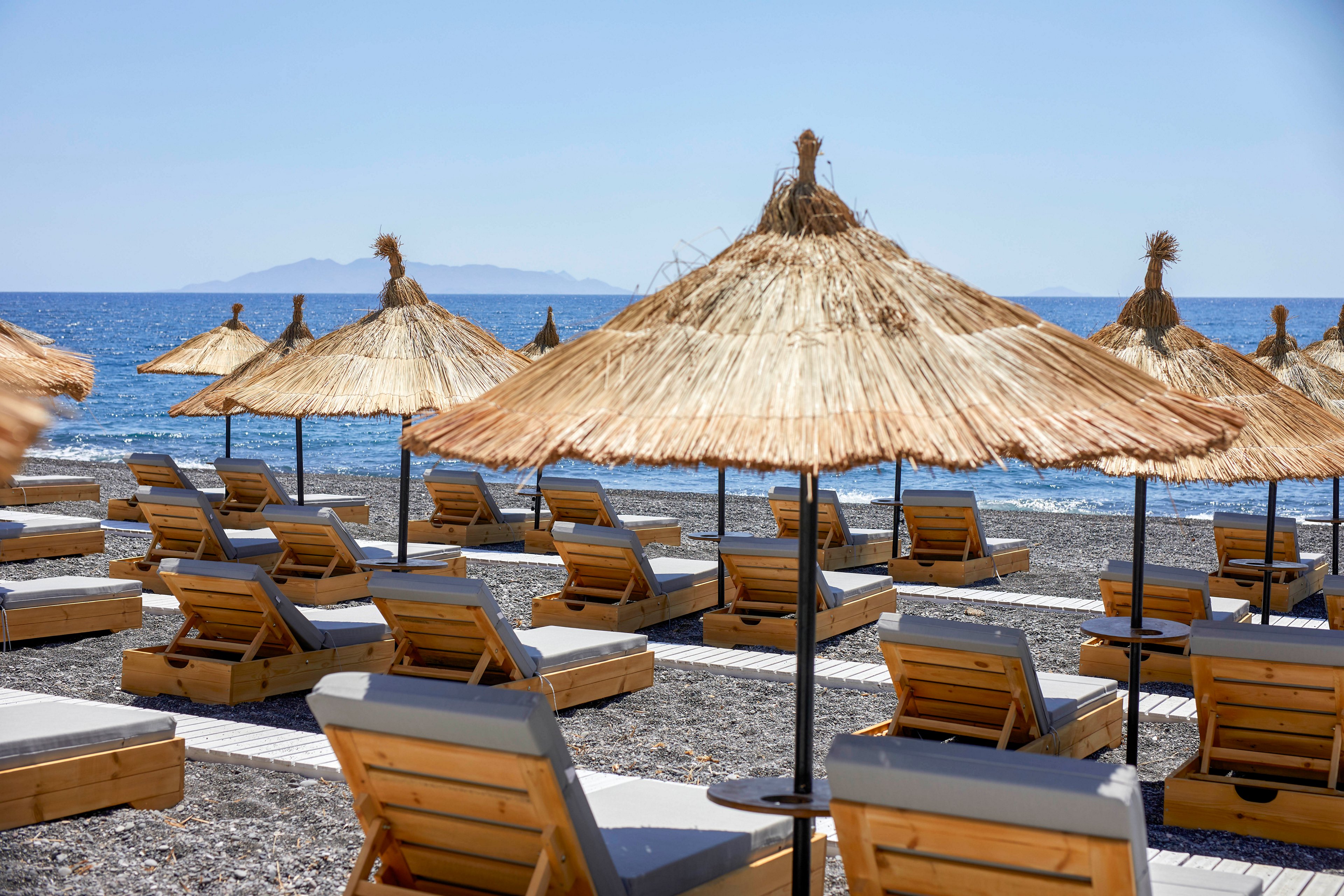 A row of wooden sun loungers with cushions sits on a pebbled beach under straw parasols. The loungers face a calm blue sea under a clear sky. No people are present, emphasizing a peaceful and quiet seaside setting. An island or distant landmass is visible on the horizon across the water.
