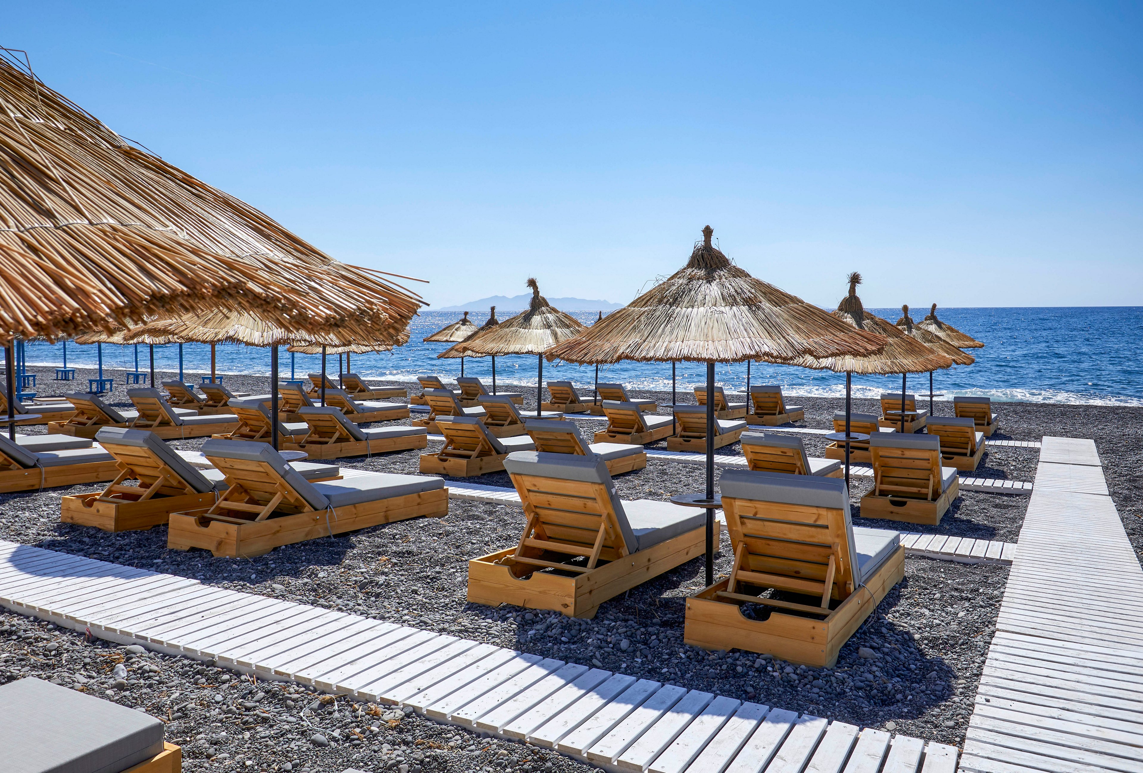 A row of wooden sun loungers with cushions is arranged on a pebble beach under straw parasols. A white wooden walkway leads between the chairs toward the shoreline. The calm blue sea stretches to the horizon under a clear sky, suggesting a warm, sunny day. The scene feels peaceful and inviting, with no people present on the beach.
