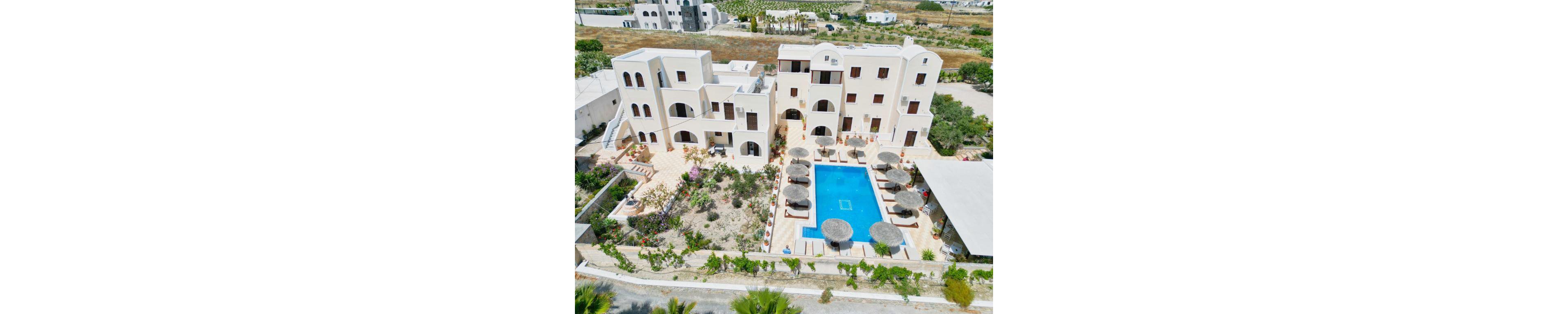 Aerial view of a Mediterranean-style villa featuring whitewashed architecture and arched windows. The property includes a central swimming pool surrounded by sun umbrellas and lounge chairs. Lush landscaping with colorful flowers and greenery enhances the serene setting. The villa is situated in a quiet area with nearby buildings visible in the background.