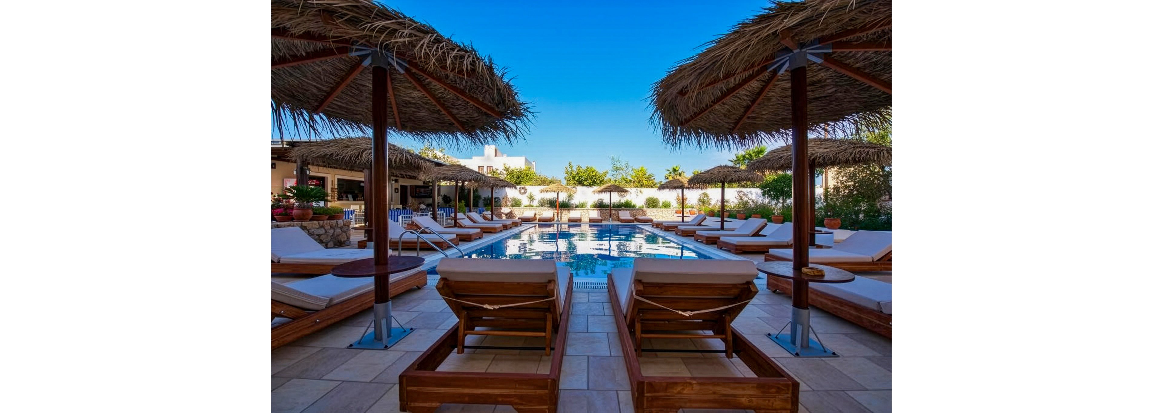 A serene outdoor pool surrounded by wooden sun loungers and straw umbrellas under a clear blue sky. The setting appears to be a resort or vacation property with lush greenery and a relaxed atmosphere. The pool area is designed for leisure and comfort, offering a tranquil escape.