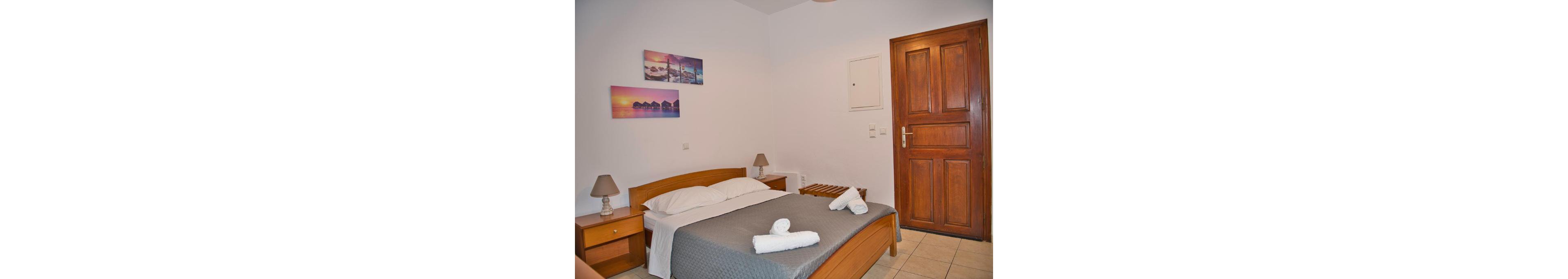 A neatly arranged hotel room featuring a wooden bed frame, bedside tables, and a door. The bed is made with white linens and a gray blanket, complemented by rolled towels. Wall art depicting serene landscapes adds a touch of color to the minimalist decor.