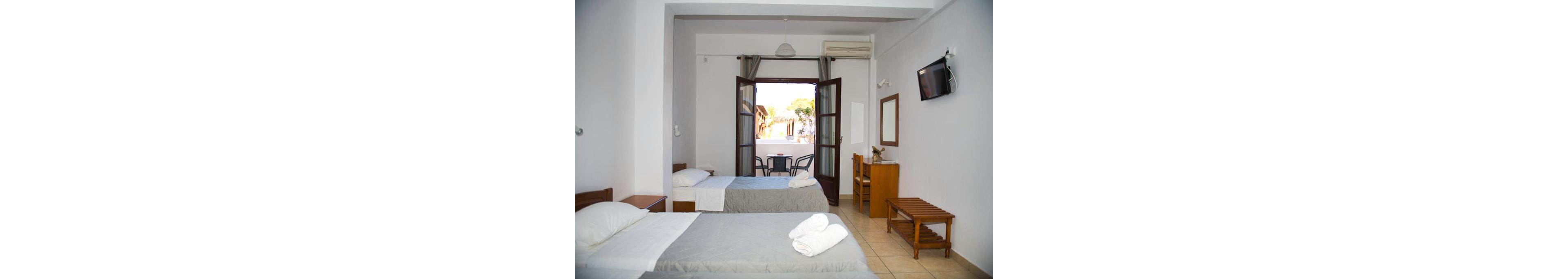 A bright and simple hotel room featuring two single beds with white and gray linens. The room includes a wooden desk, chair, and wall-mounted TV. French doors open to a balcony with outdoor seating and a view of trees and clear skies.