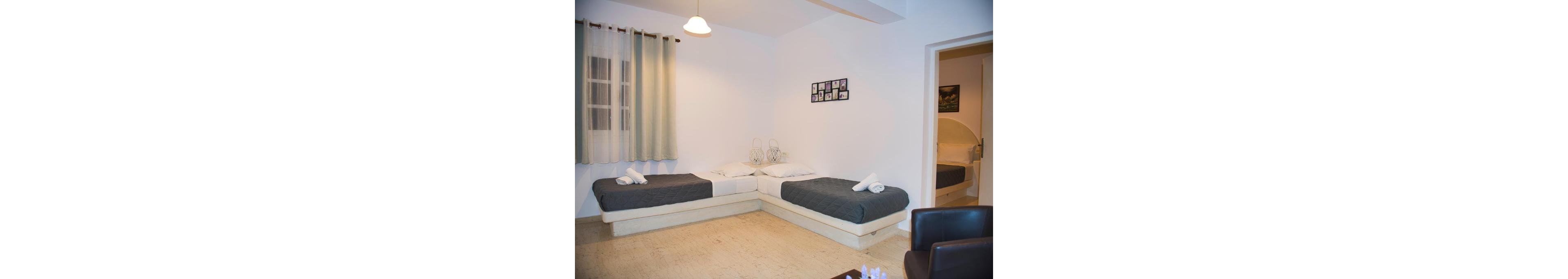 A well-lit room featuring two twin beds with dark gray bedding and neatly folded towels. The space includes minimalistic decor, a wall-mounted photo frame, and a view of an adjoining room. The ambiance is clean and inviting, with soft lighting and neutral tones.