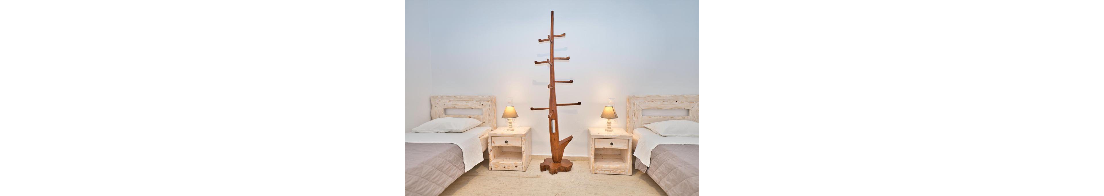 A clean and minimalist twin bedroom featuring two single beds with beige bedding and rustic wooden headboards. The room is accented by a unique wooden tree-shaped coat rack placed centrally between the beds. Warm lighting from bedside lamps adds a cozy ambiance to the neutral-toned space.