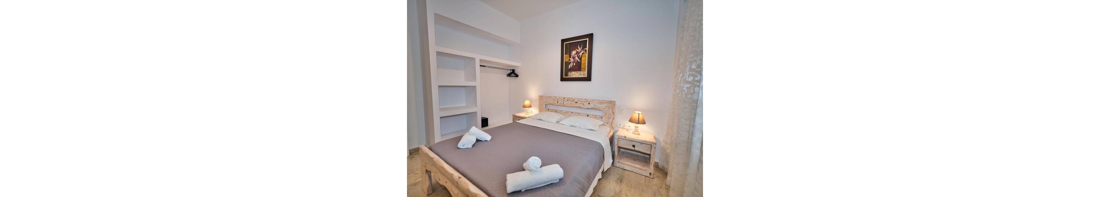 A neatly arranged bedroom featuring a wooden bed frame and matching nightstands. The setting includes soft lighting from bedside lamps and folded towels placed on the bed. The decor is minimalistic with a framed floral artwork on the wall and an open closet space visible in the background.