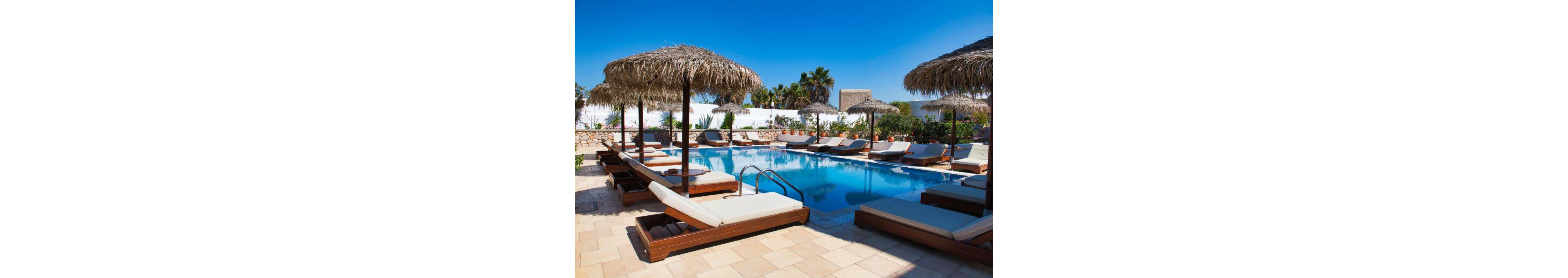 A serene outdoor pool surrounded by wooden sun loungers and straw umbrellas, set in a sunny, tropical environment. The pool area is bordered by lush greenery and white walls, creating a relaxing vacation vibe. Clear blue skies enhance the tranquil atmosphere, perfect for leisure and relaxation.