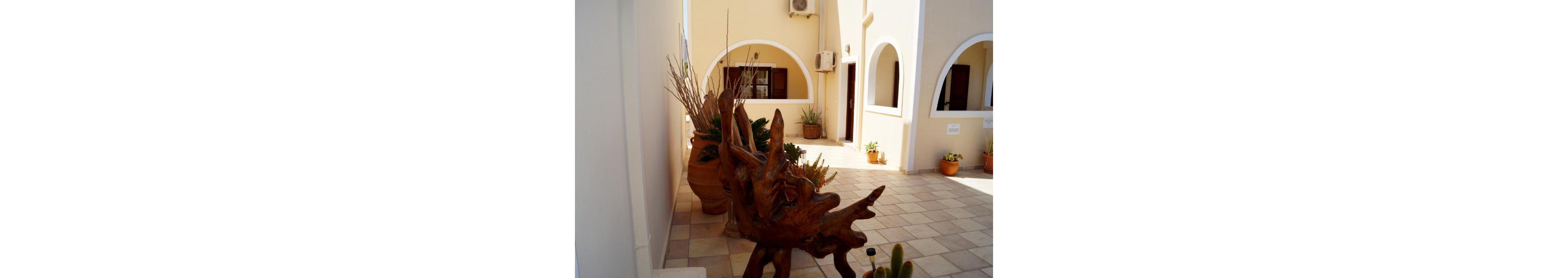 A serene Mediterranean-style courtyard featuring rustic wooden sculptures and terracotta pots. The setting includes tiled flooring, arched windows, and walls painted in neutral tones. Succulent plants and cacti add a touch of greenery to the space, creating a warm and inviting atmosphere.