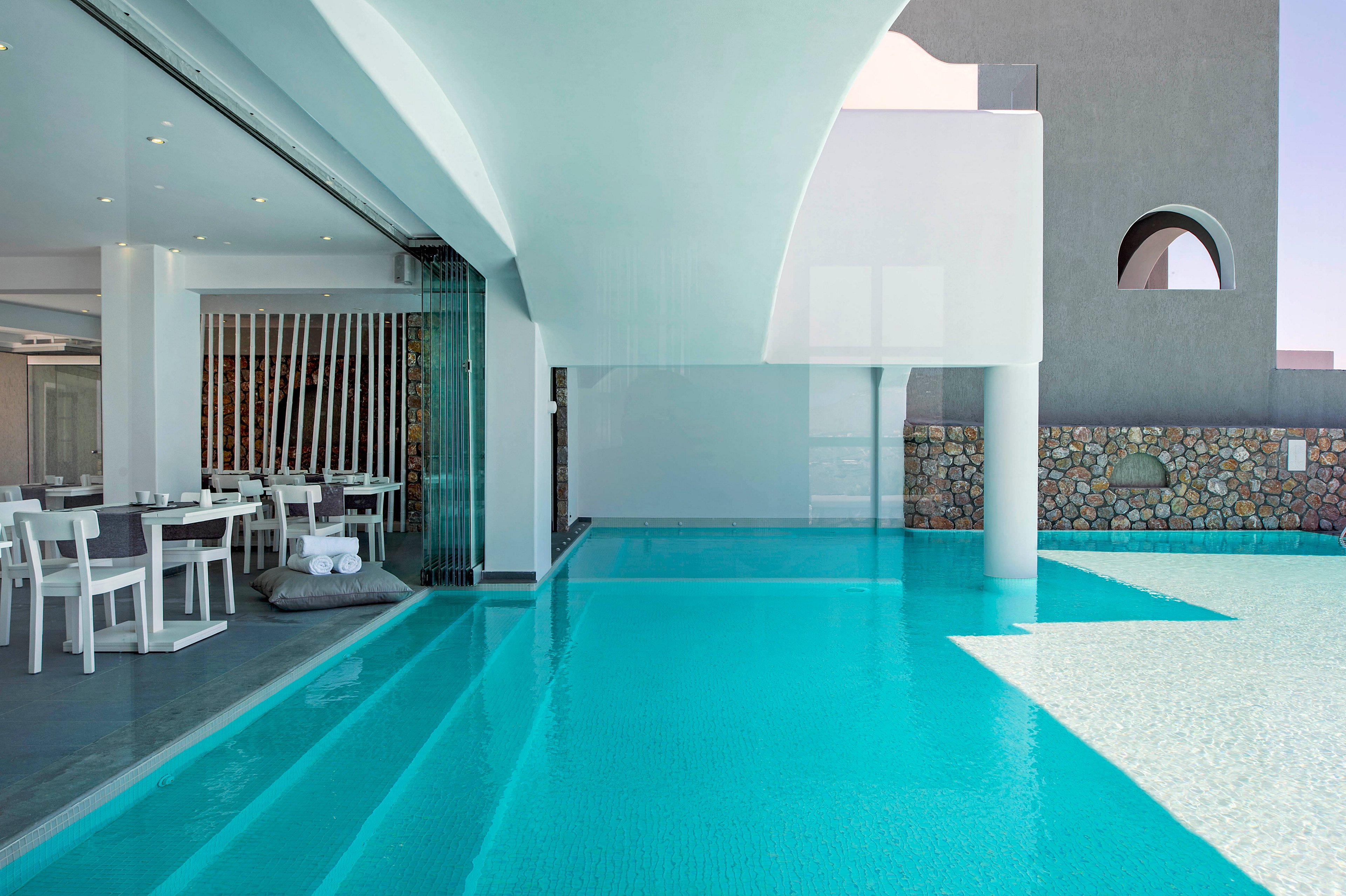 A contemporary resort features a sleek poolside dining area with white chairs and tables. The turquoise swimming pool extends partially indoors, reflecting the minimalist architecture and clean lines. Natural stone accents and geometric windows add visual interest to the serene setting. The overall mood is tranquil and inviting, with a cool color palette dominating the scene.