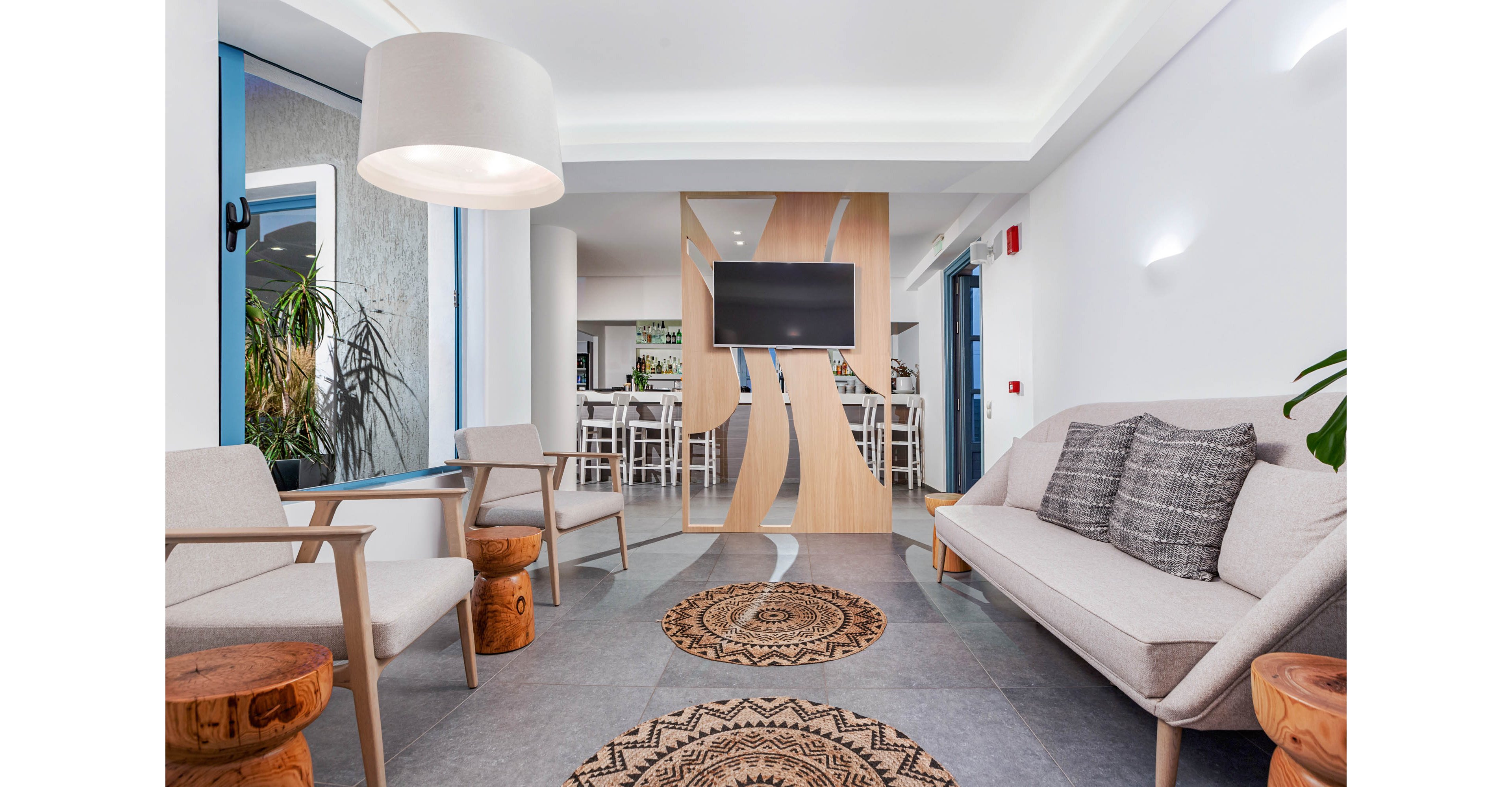 A contemporary lounge features light-colored armchairs and a sofa with textured pillows, arranged around wooden accent tables. The space is defined by a decorative wall with a mounted flat-screen TV and geometric patterns. Natural light enters through large windows, and the overall palette is neutral with touches of wood and gray. The setting appears calm and inviting, suitable for waiting or casual meetings. No visible text or numbers are present in the image.