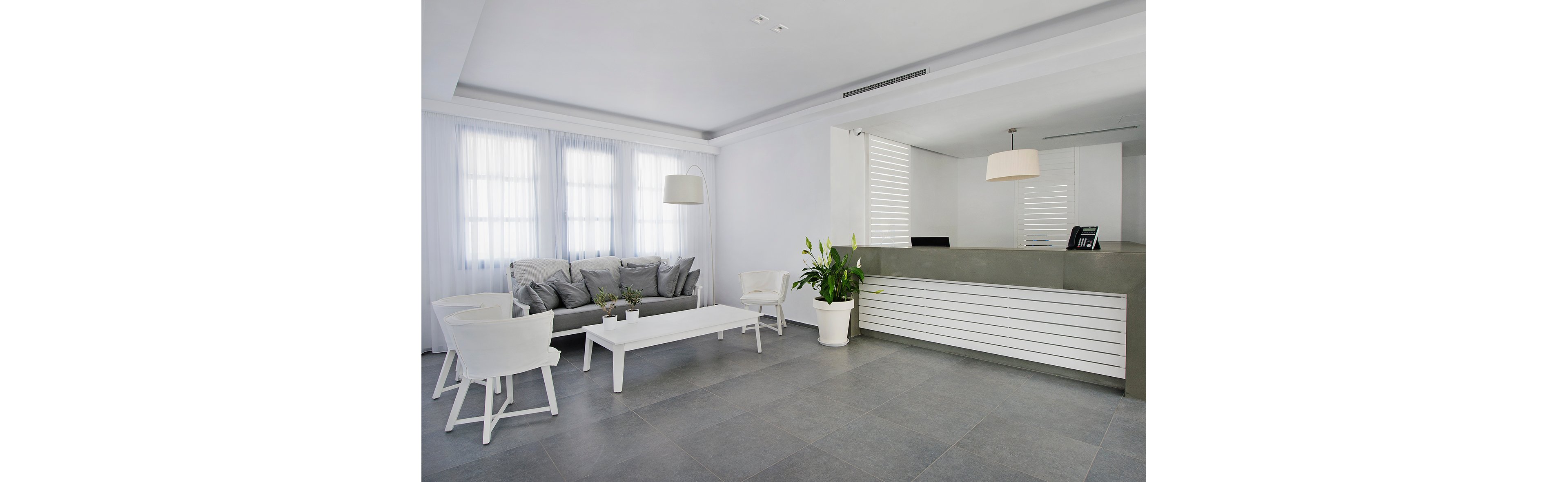 A contemporary hotel lobby features a sleek white reception desk and a cozy seating area with a gray sofa and white chairs. Large windows with sheer curtains allow natural light to fill the space, highlighting the minimalist design. A potted plant adds a touch of greenery, while pendant lights provide soft illumination. The overall palette is neutral, creating a calm and inviting atmosphere.