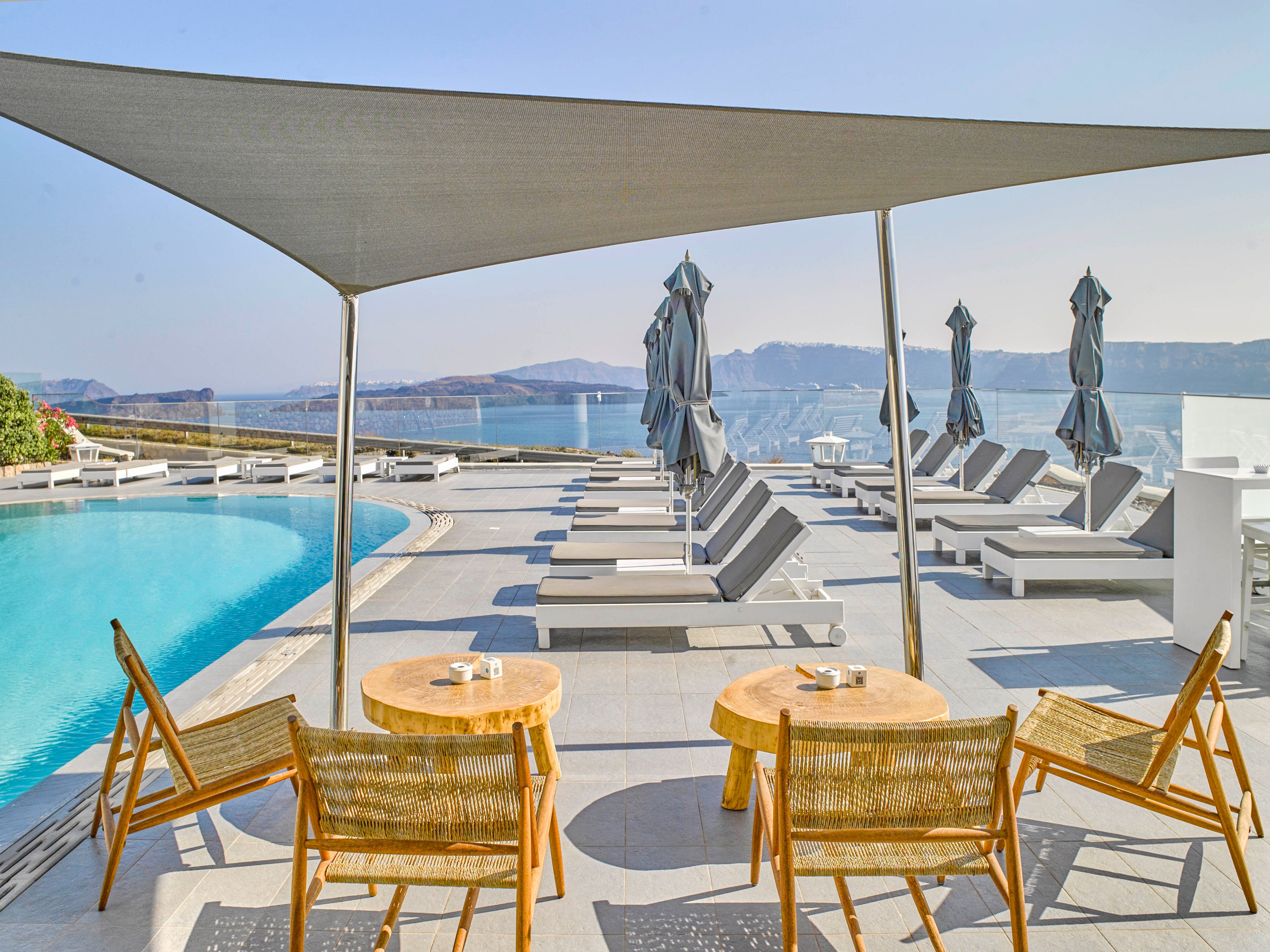 A contemporary resort pool area features wooden tables and chairs under a shade canopy, overlooking a serene infinity pool. Multiple sun loungers with closed umbrellas line the poolside, facing a stunning ocean and mountain backdrop. The setting is bright and tranquil, with a minimalist design and neutral color palette. The atmosphere suggests relaxation and luxury in a coastal environment.