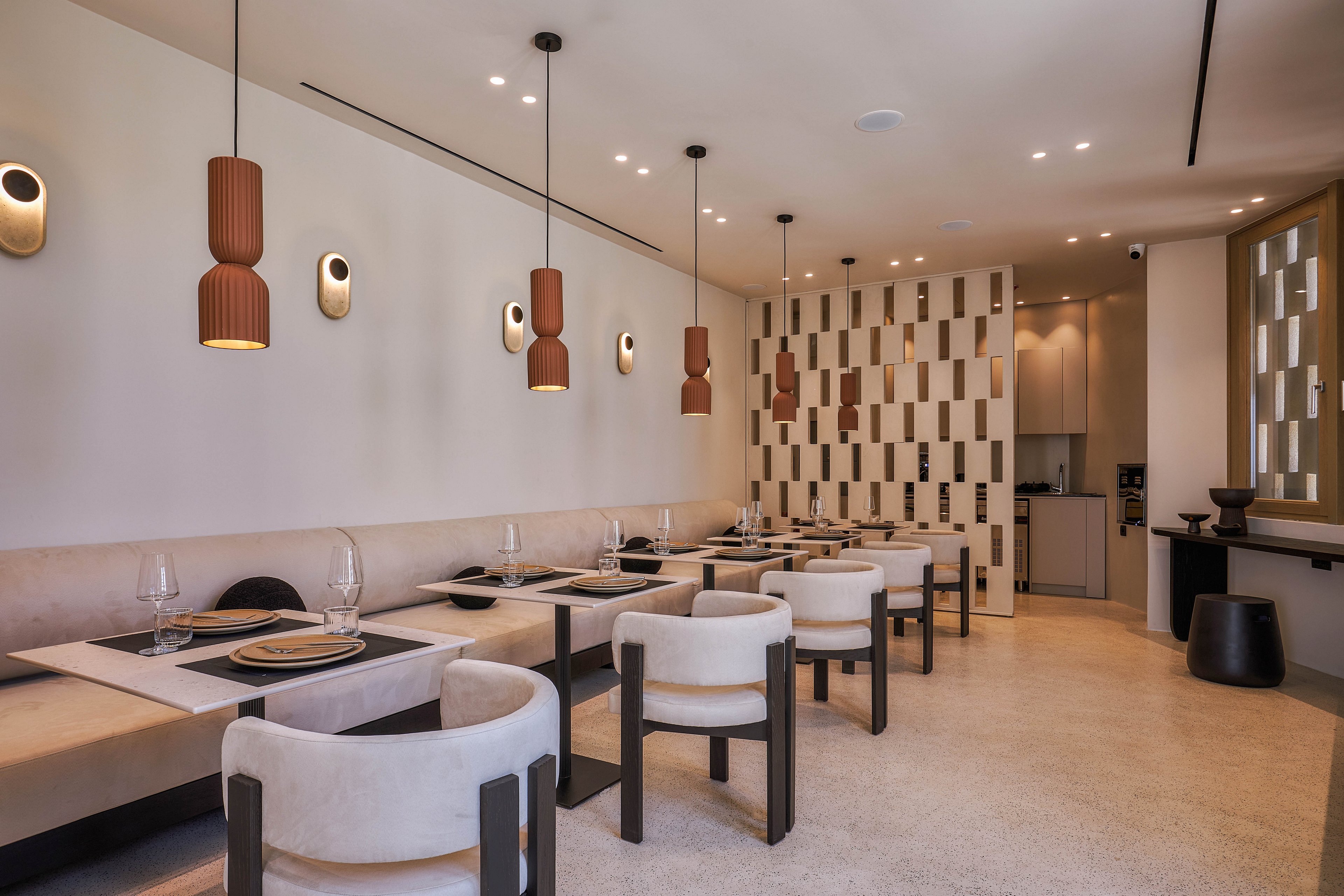 A contemporary restaurant interior features neatly arranged tables and cushioned bench seating along a light-colored wall. Pendant lights and wall sconces provide warm, ambient illumination, highlighting the minimalist decor. Neutral tones dominate the space, with beige, cream, and brown surfaces creating a calm atmosphere. A decorative partition and an open view to the service area add depth and visual interest to the dining room.