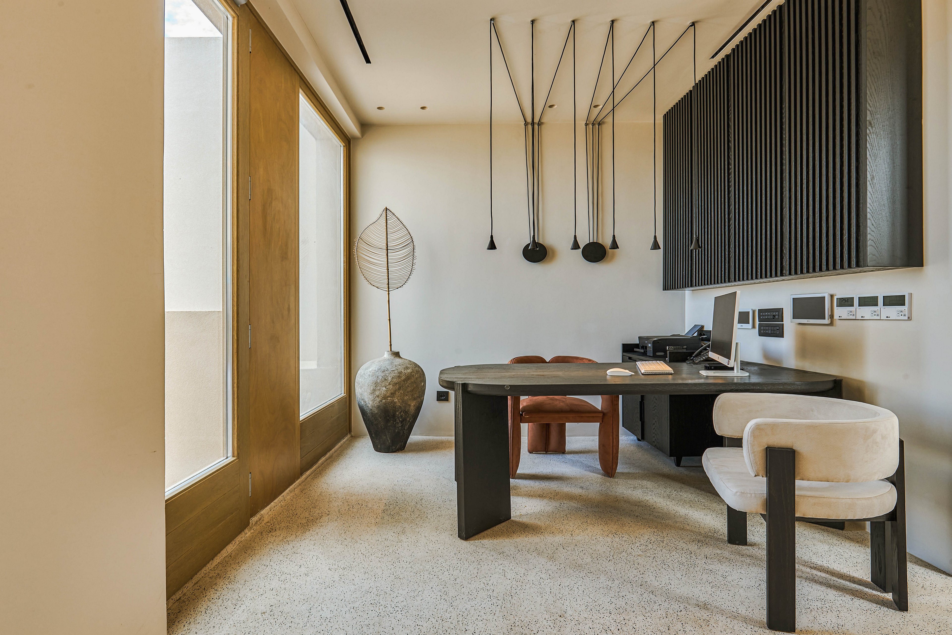 A contemporary home office features a sleek black desk with a computer and accessories arranged neatly on top. The room includes two modern upholstered chairs, one behind the desk and one in front, set on a light carpeted floor. Large windows on the left bring in natural light, while sculptural lighting fixtures and a tall decorative vase add artistic interest. Dark wall panels and clean lines emphasize a minimalist, neutral-toned interior design.
