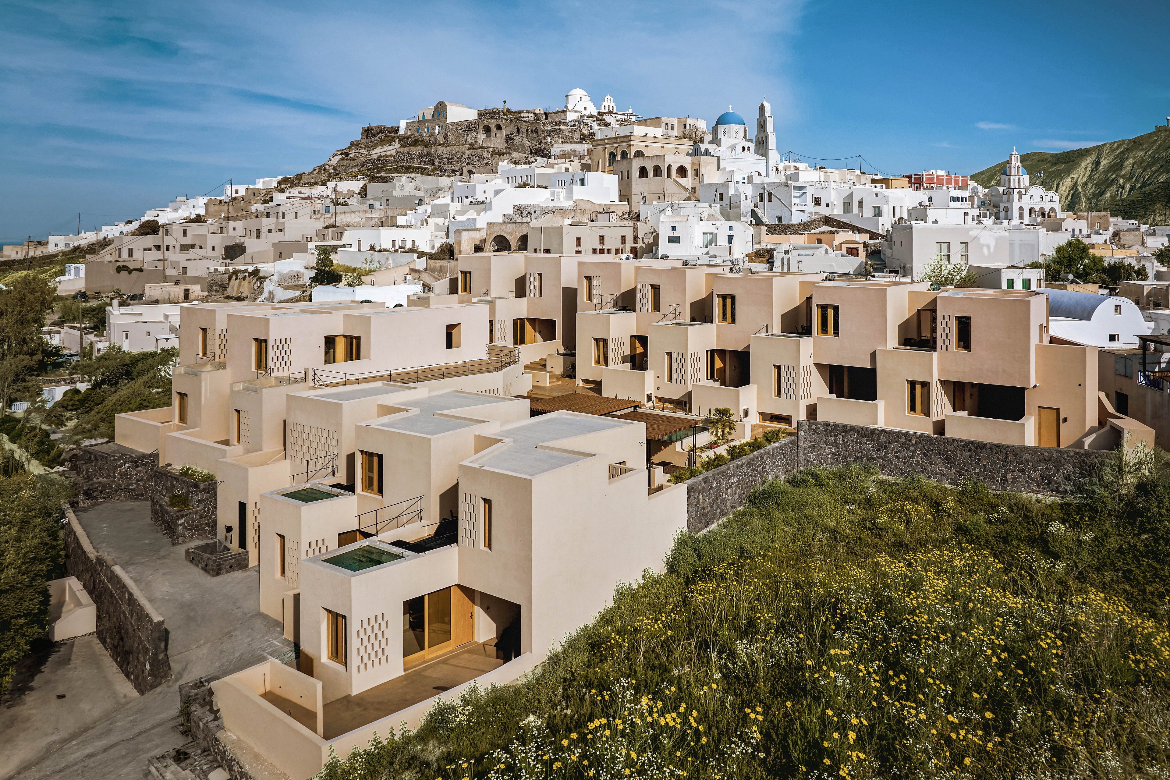 A cluster of modern beige apartment buildings sits on a hillside, overlooking a traditional whitewashed village. The architecture features clean lines, terraces, and balconies that step down the slope. In the foreground, wild greenery and yellow flowers contrast with the angular structures. The background reveals densely packed houses and a blue sky, suggesting a sunny coastal or island setting.