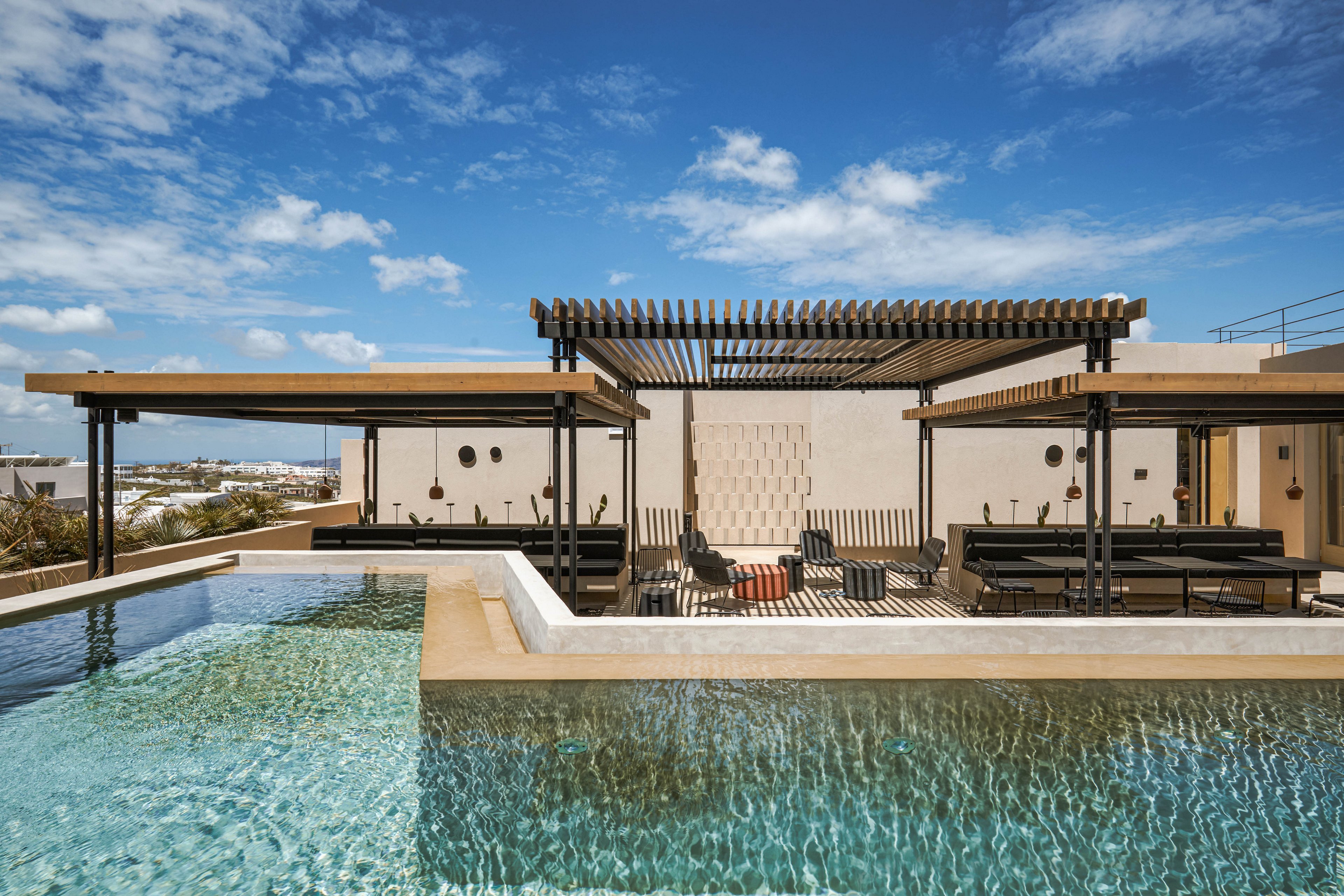 A contemporary rooftop infinity pool stretches across the foreground, reflecting the blue sky and scattered clouds. Behind the pool, a shaded lounge area with dark pergolas and minimalist seating creates an inviting outdoor retreat. The architecture features clean lines, neutral beige walls, and geometric details that emphasize a modern design aesthetic. In the distance, a low urban or coastal landscape is visible under bright daylight, enhancing the open, airy atmosphere.