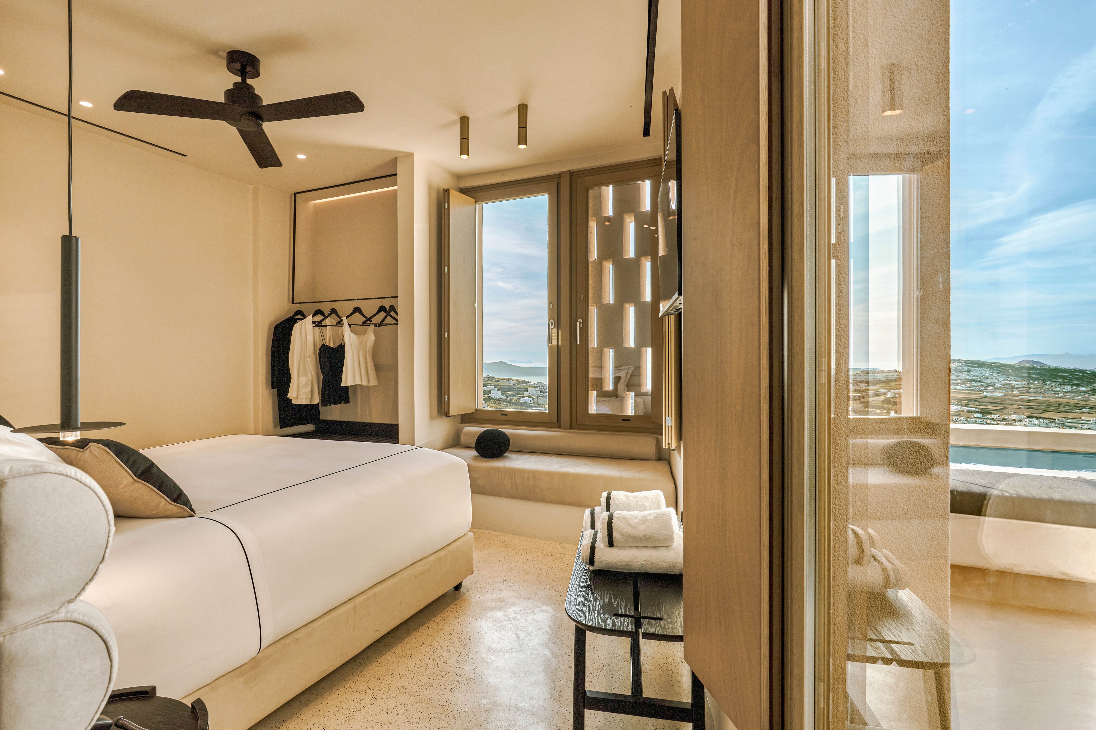 A contemporary hotel bedroom features a large bed with crisp white linens and subtle black accents. The room includes an open wardrobe with neatly hung clothing and a bench with folded towels. Warm beige tones, natural light, and sleek fixtures create a minimalist, luxurious atmosphere. Large windows reveal an expansive coastal or seaside view in the distance.