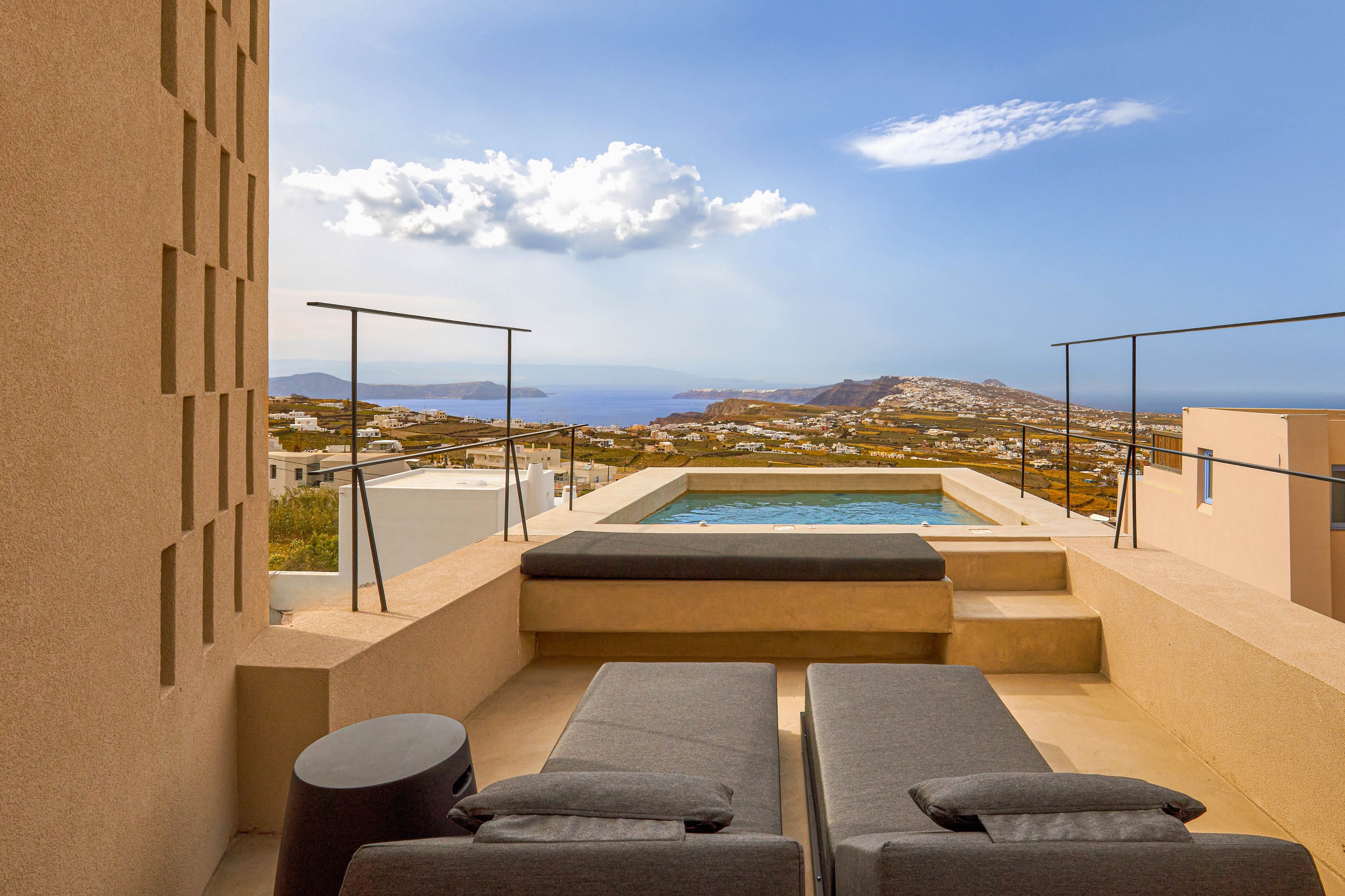 A contemporary terrace features two cushioned lounge chairs facing a small rectangular plunge pool. The space is framed by minimalist beige walls and simple metal railings. Beyond the pool, there is a panoramic view of a coastal town and the distant sea under a partly cloudy sky. The scene suggests a quiet, luxurious vacation setting ideal for relaxation and sunbathing.